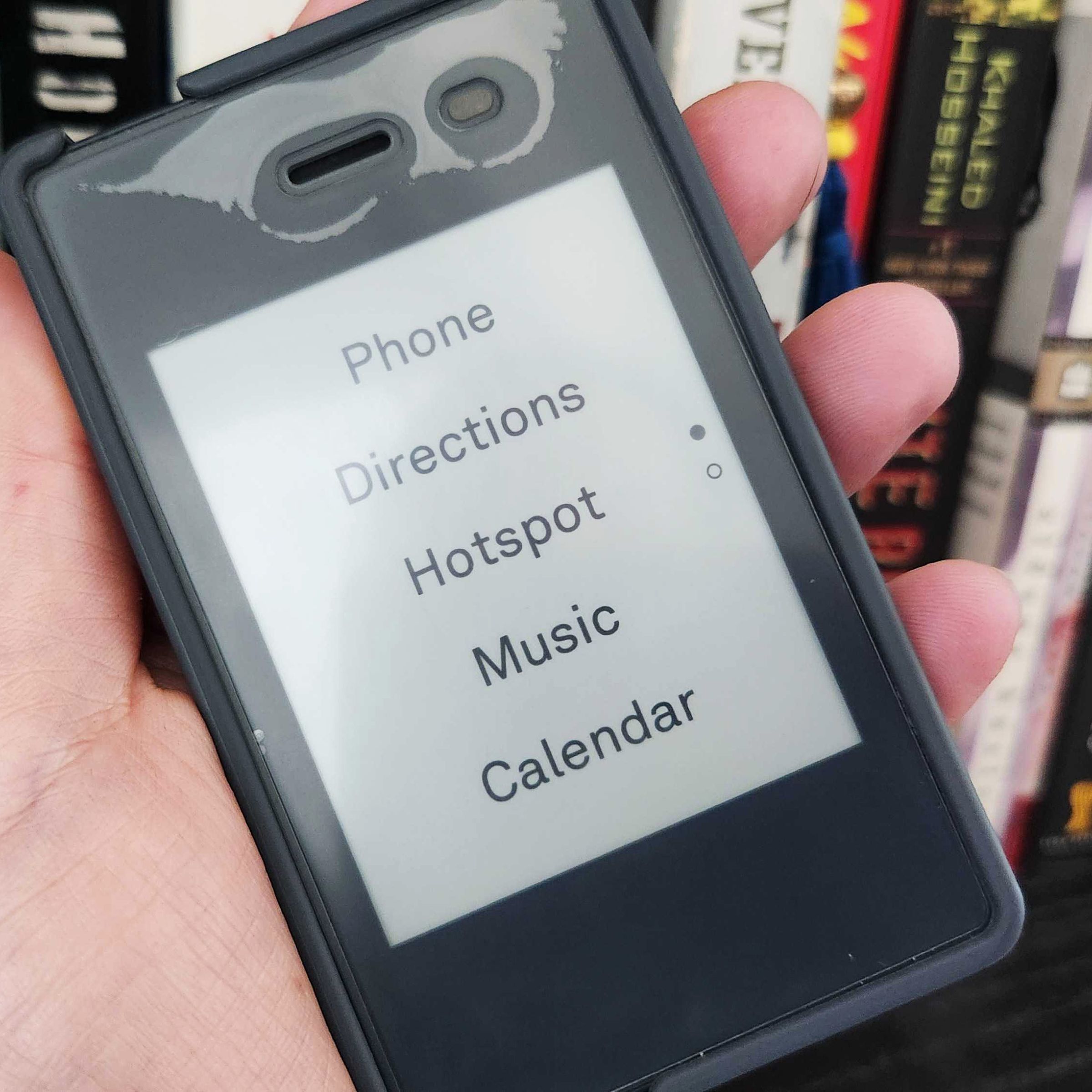 A small cellphone held in a person’s hand with books in the background.