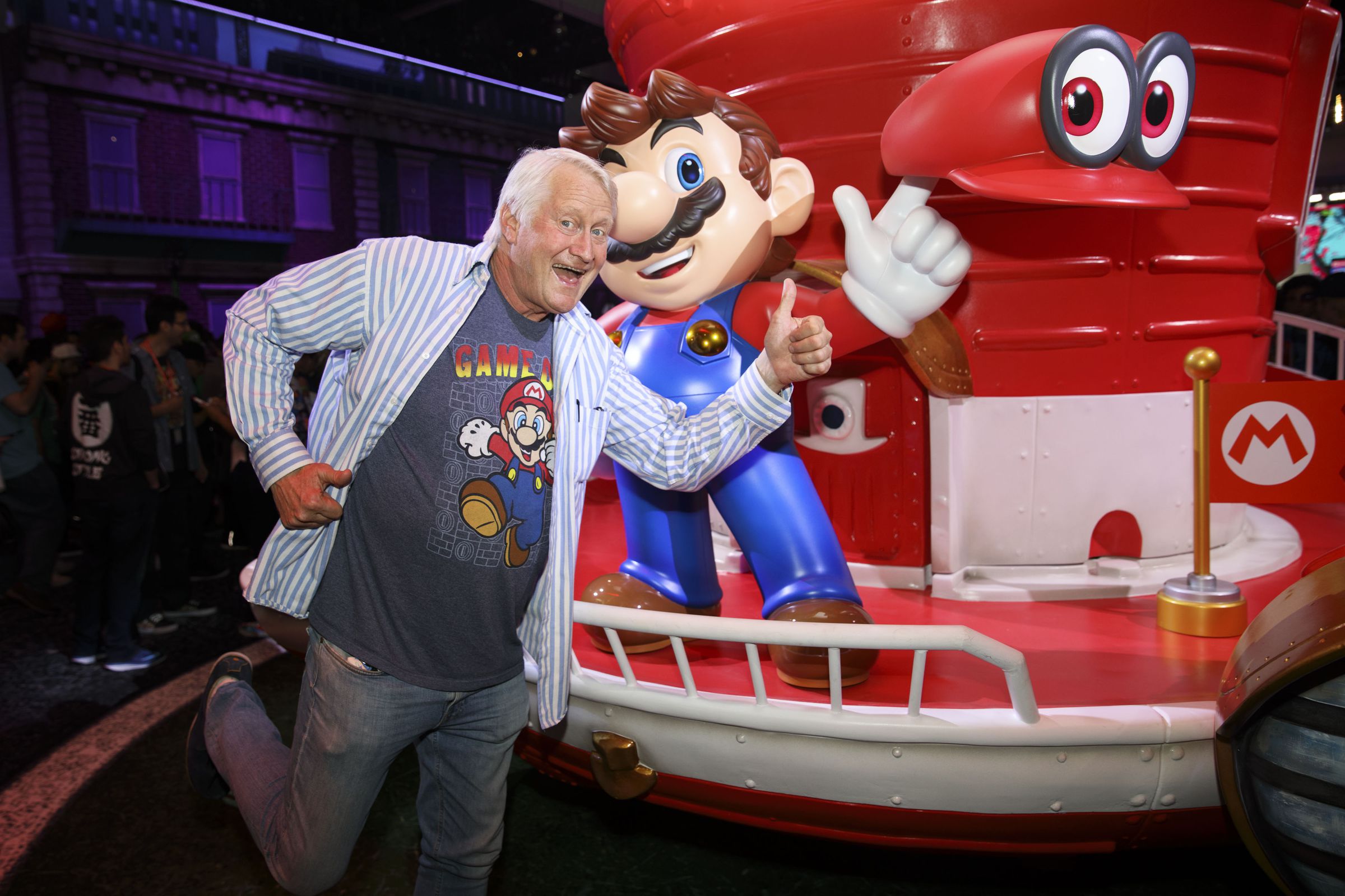 A photo of Mario voice actor Charles Martinet at E3 2017 standing next to a statue of Mario promoting Super Mario Galaxy