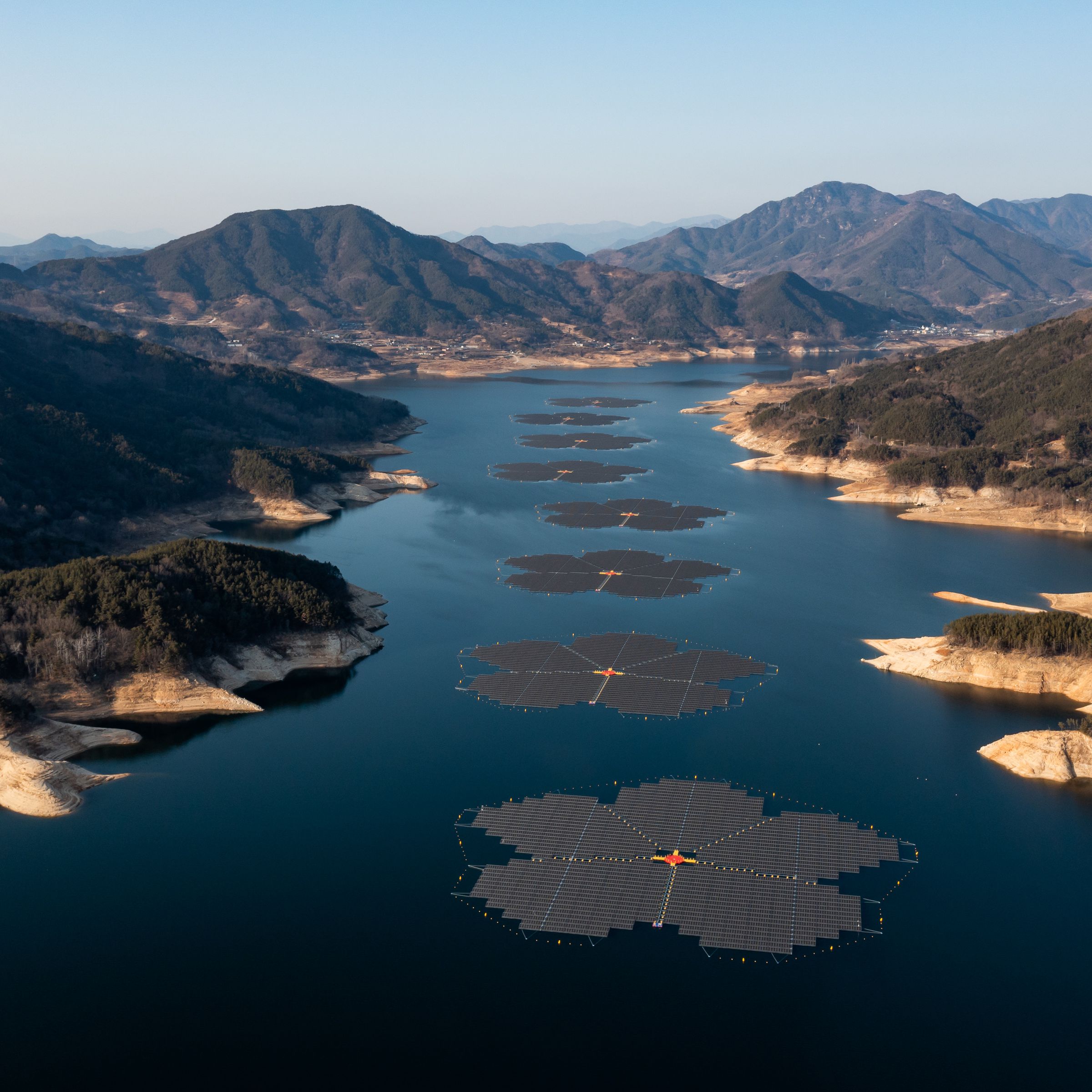 Multiple solar arrays in the shape of flowers are lined up on top of a lake between hills and mountains.