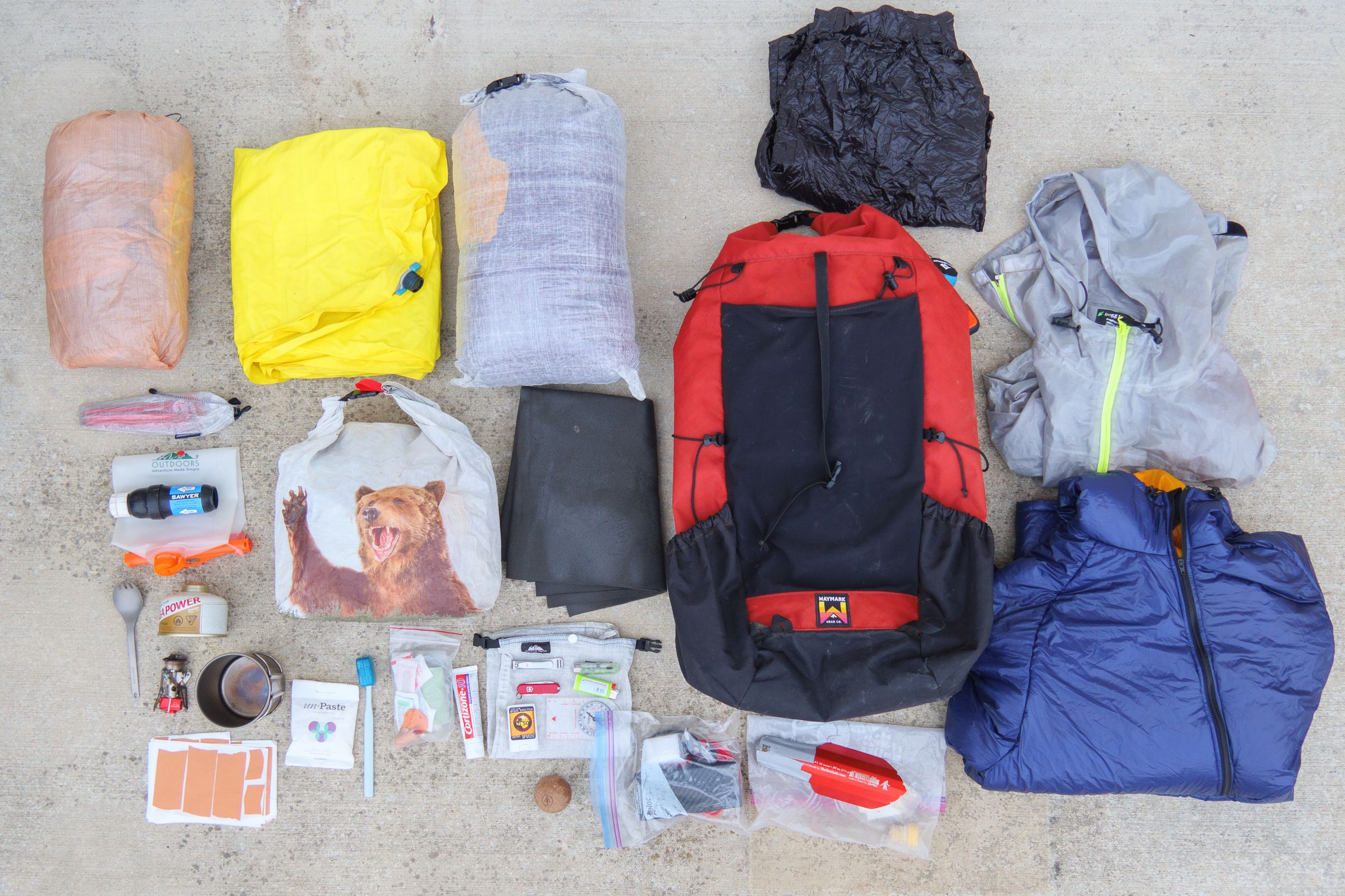 Photo of backpacking gear. From top left to bottom right: A tent, sleeping pad, quilt, wind pants, rain jacket, tent stakes, a water bag and filter, a food bag, a small foam mat, a backpack, a rain jacket, a spork, a gas canister, a mug, some leukotape strips, toothpaste tablets, a toothbrush, a first aid kit, lotion, a bag with nail clippers, a flashlight, swiss army knife, lighter, sunscreen, and compass on it, a cork ball, an electronics bag, a trowel, and a puffy jacket.