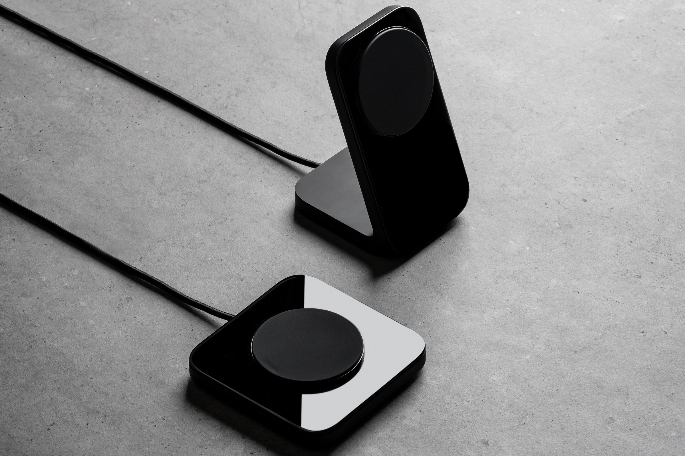A marketing image of the Nomad Stand and Nomad Base magnetic wireless chargers, in black, sitting beside each other on a gray surface.
