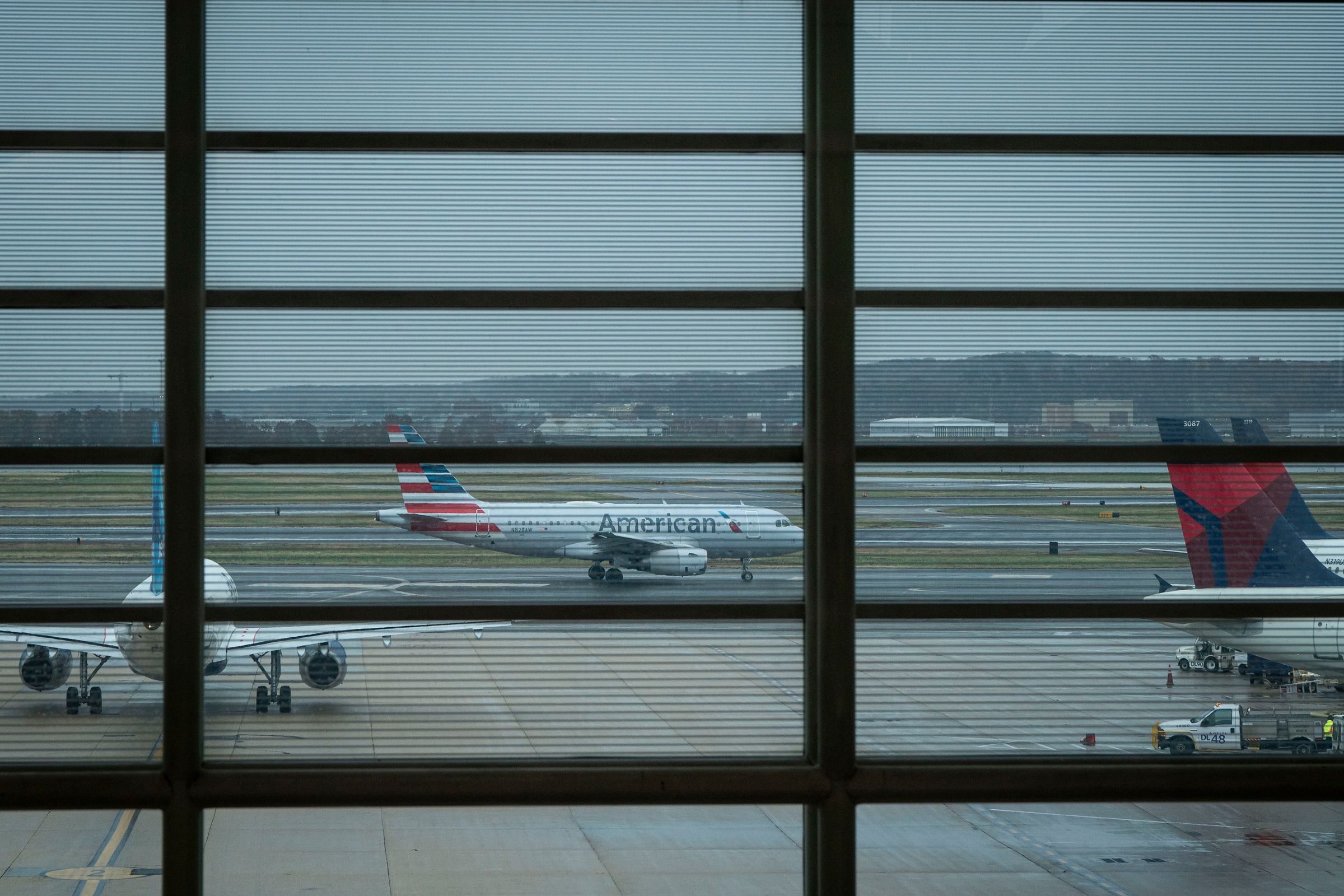 Several planes sitting on a runway seen through an airport window. A plane with the American Airlines logo can be seen in the center of the photo between window panes.