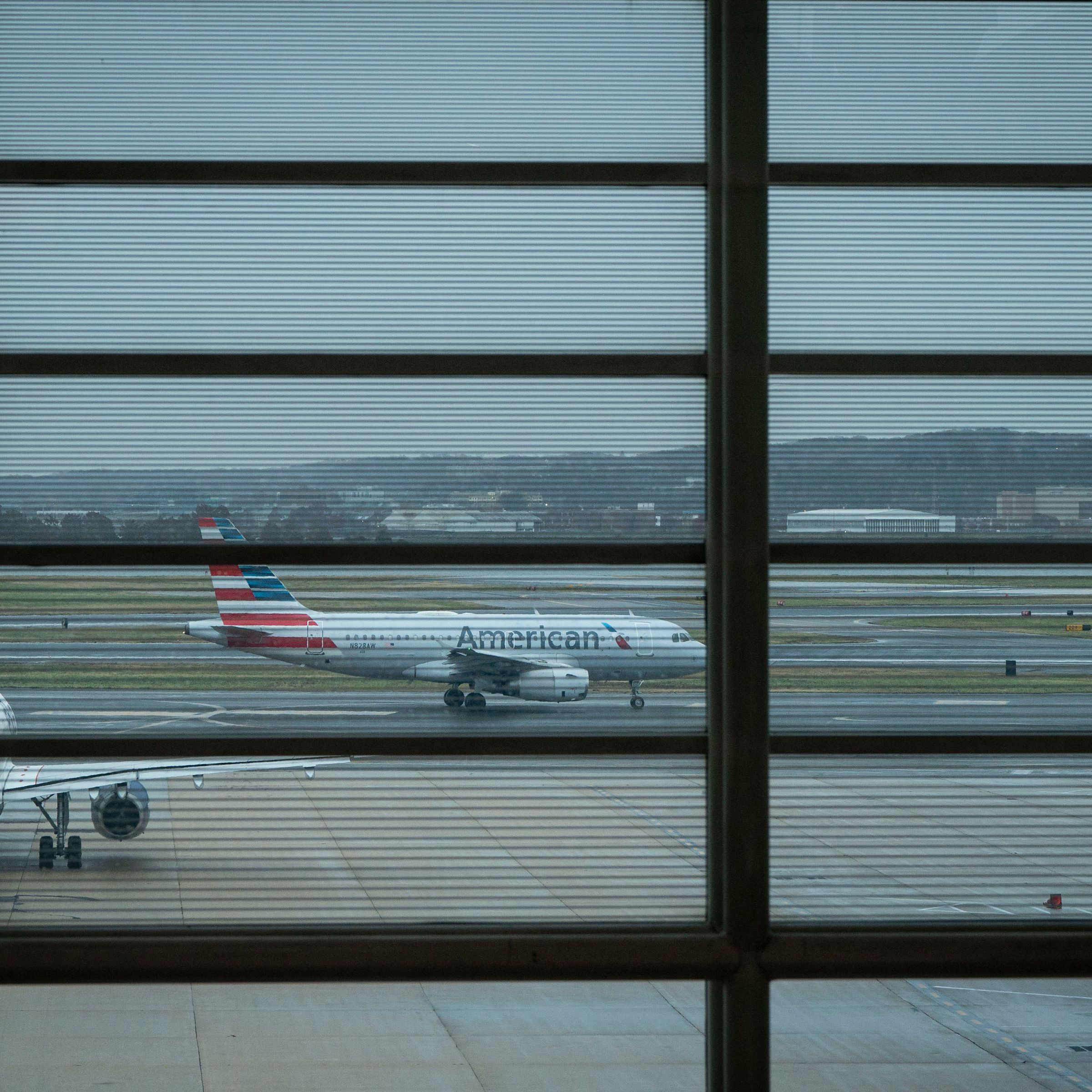 Several planes sitting on a runway seen through an airport window. A plane with the American Airlines logo can be seen in the center of the photo between window panes.