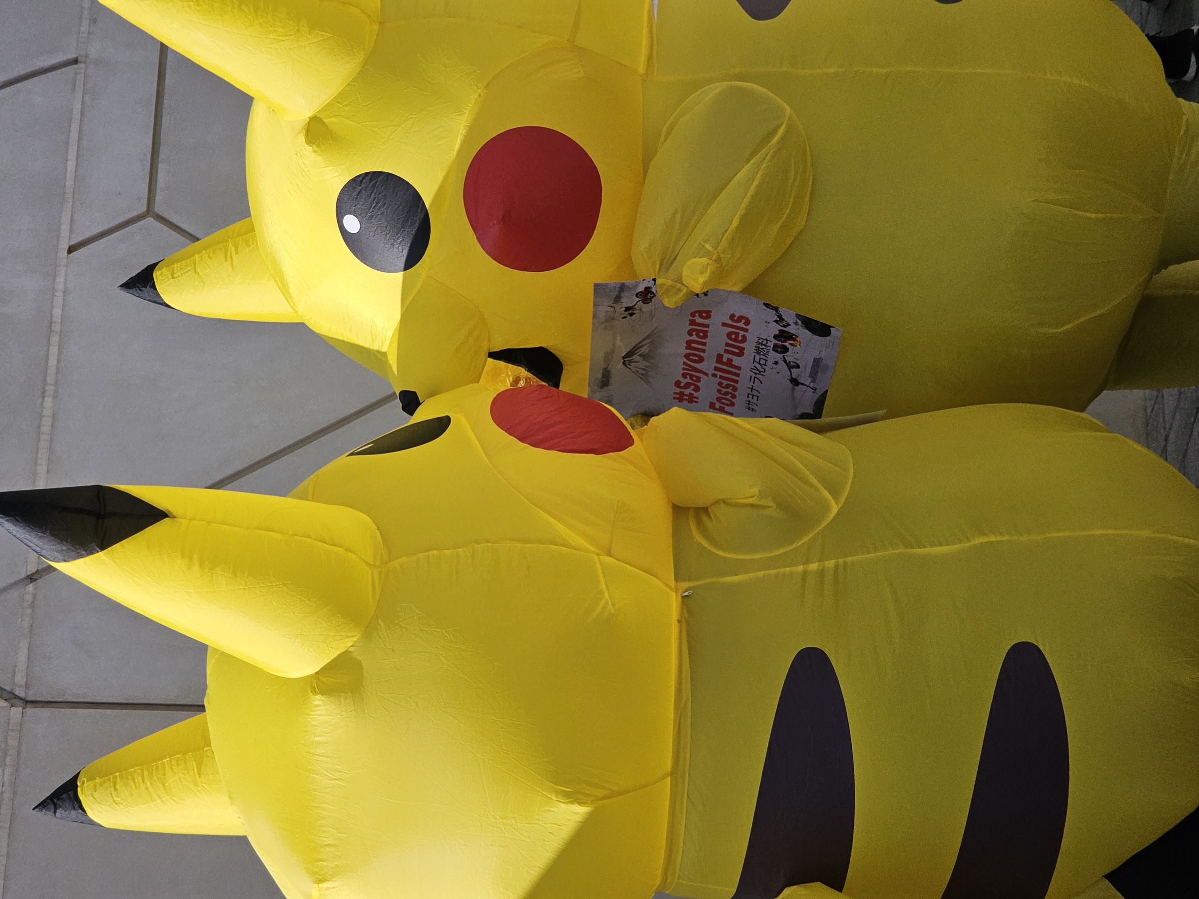 Two people wearing inflatable Pikachu costumes face each other. One holds a sign that says “#Sayonara FossilFuels”.