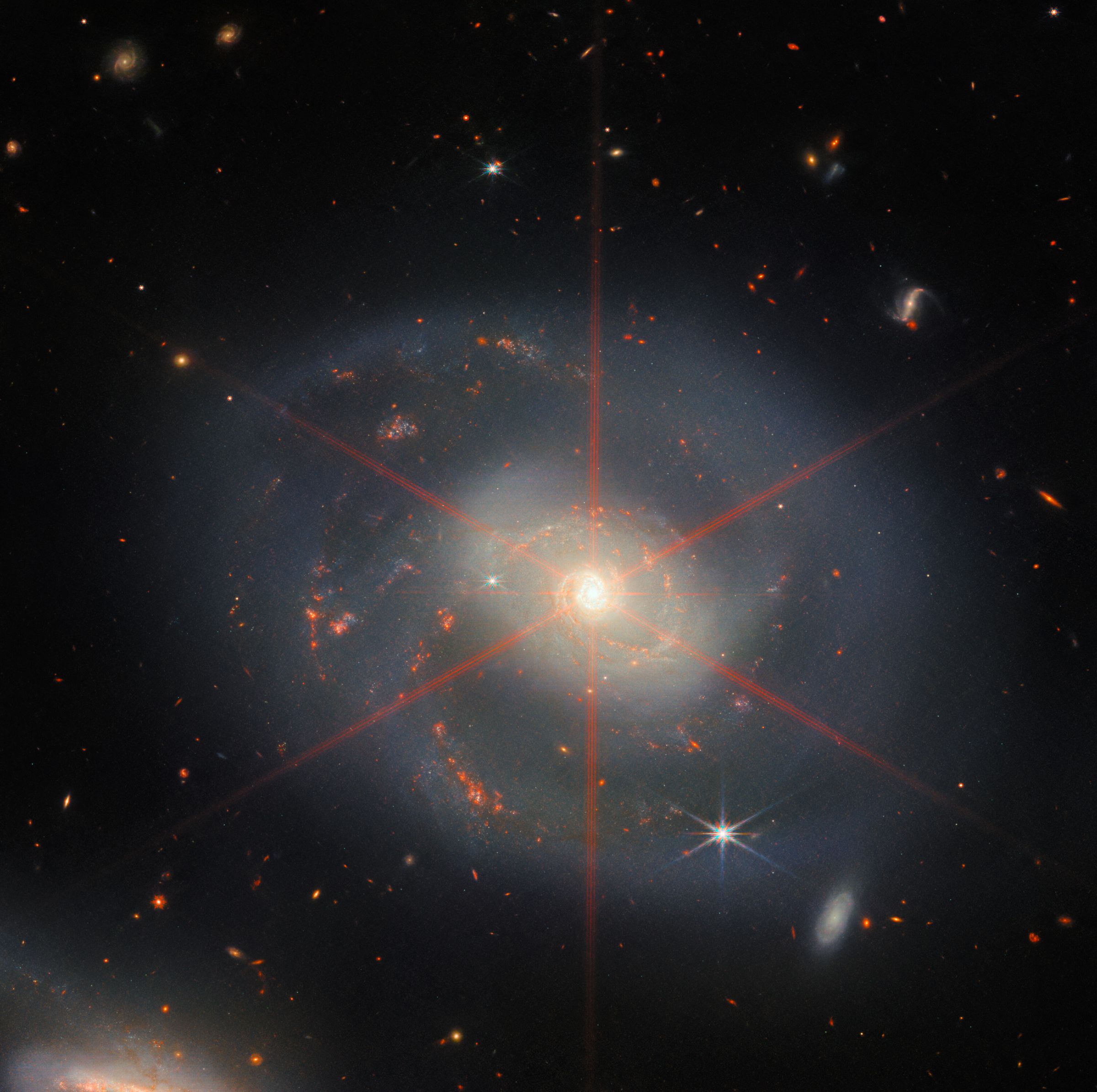 This image shows a spiral galaxy dominated by a bright central region. The galaxy has a blue-purple hue, with orangy-red regions filled with stars. Also visible is a large diffraction spike, appearing as a pattern of stars over the central region of the galaxy.Many stars and galaxies fill the background