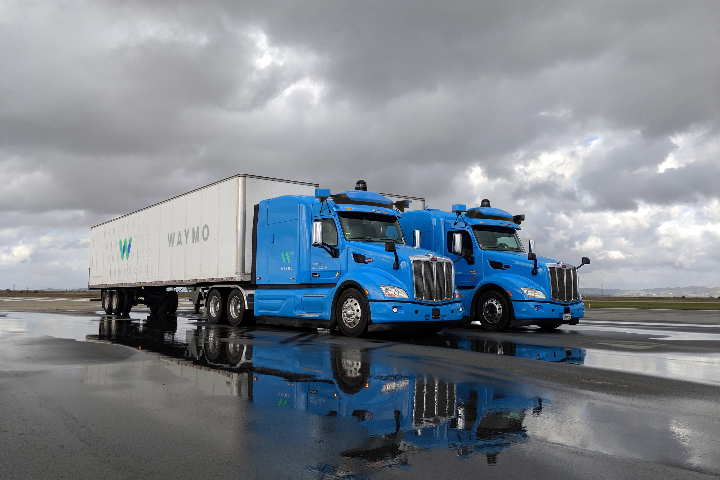 Two blue Waymo autonomous trucks next to one another on a cloudy, rainy day.