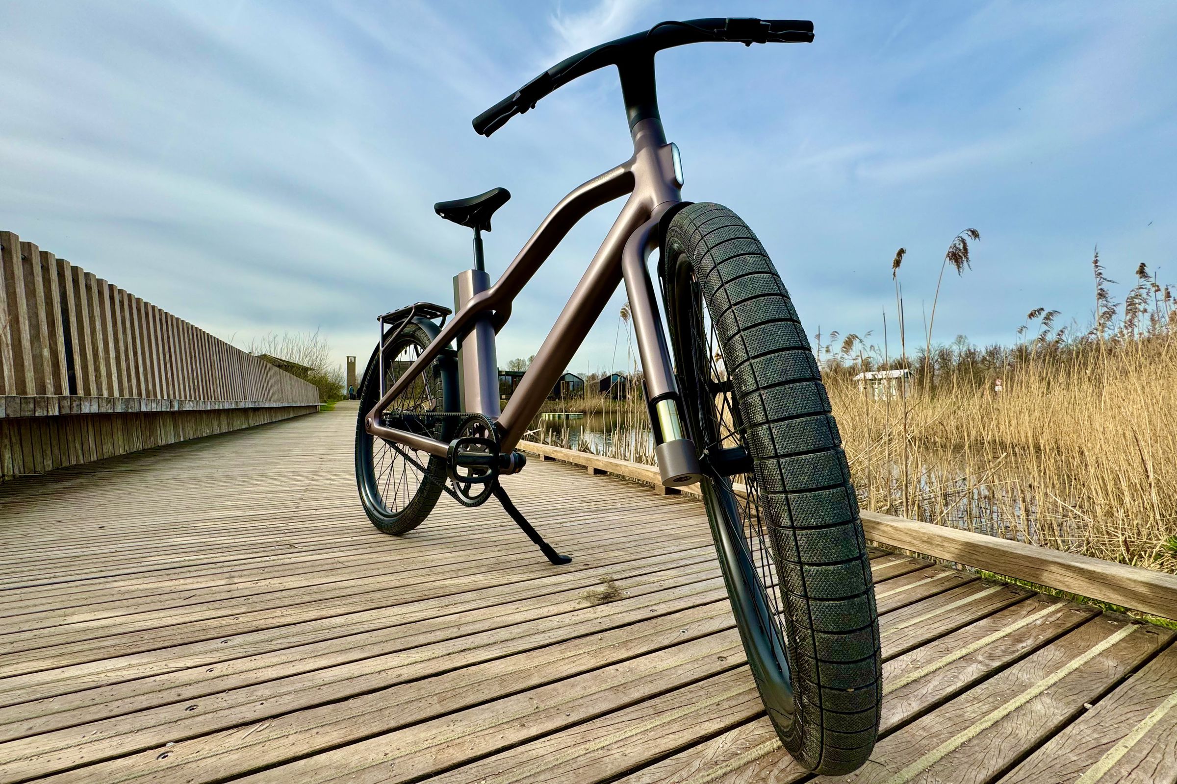 A menacing-looking Cowboy Cross e-bike sits on a wooden bridge that traverses a pond with tall grass surrounding it.