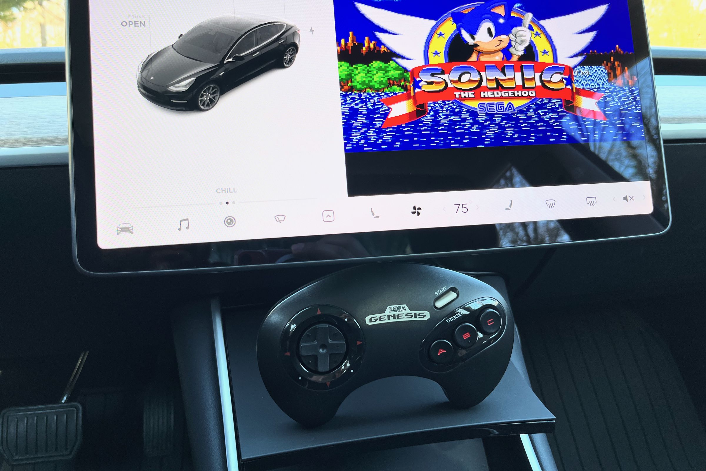 The Tesla Model 3 interior is displayed with a Sega Genesis game controller sitting under the infotainment screen with a window on the screen showing the game Sonic 1.