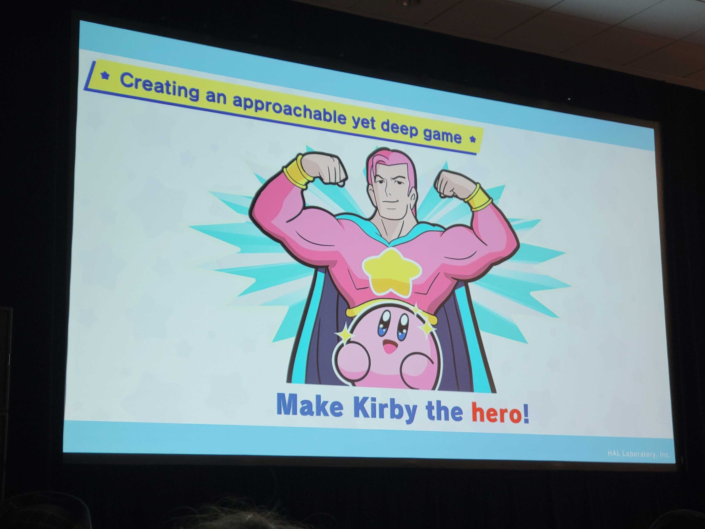Photo taken from “The Many Dimensions of Kirby” presentation at GDC 2023 featuring Kirby standing in front of a buff cartoon man wearing a superhero costume