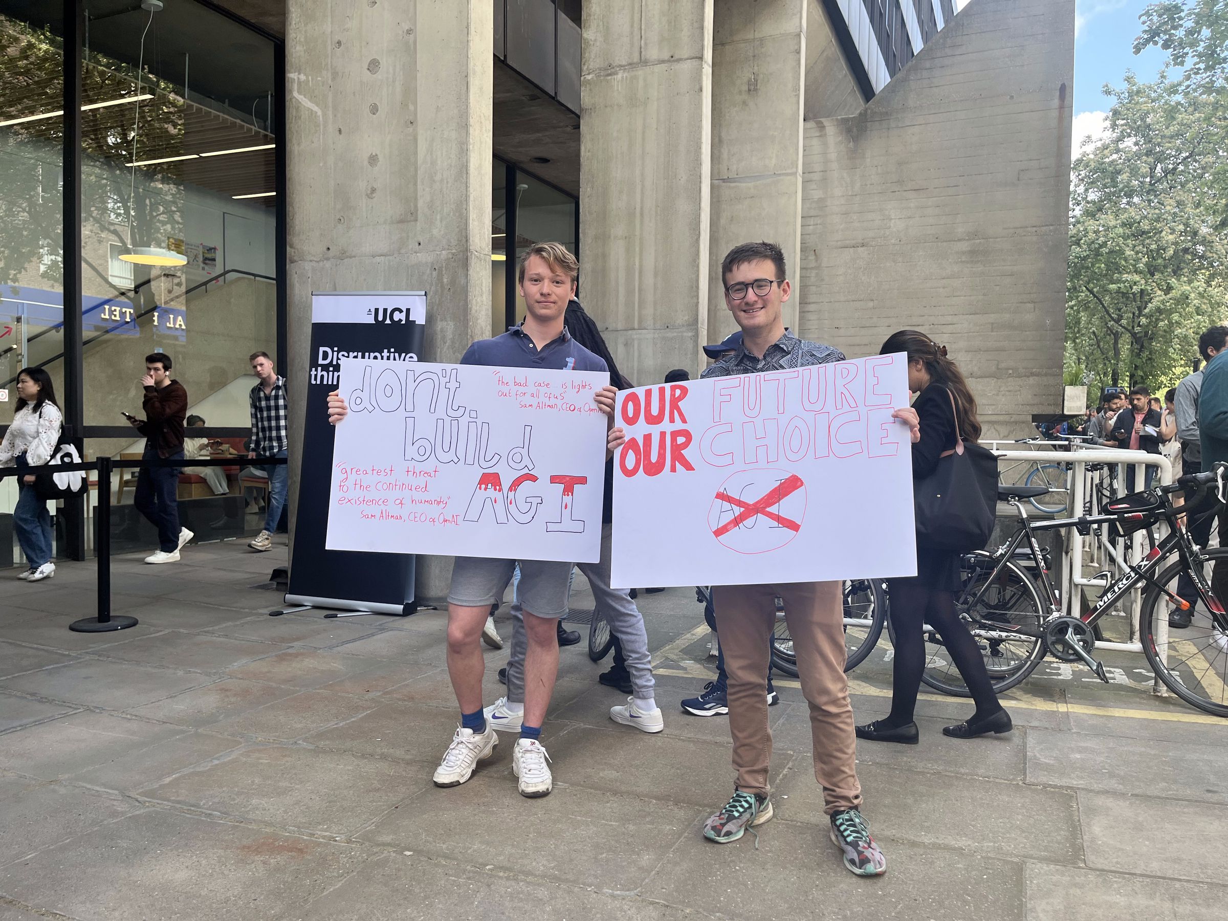 Two young men hold signs in front of a queue saying “don’t build AGI” and “our future, our choice.” 