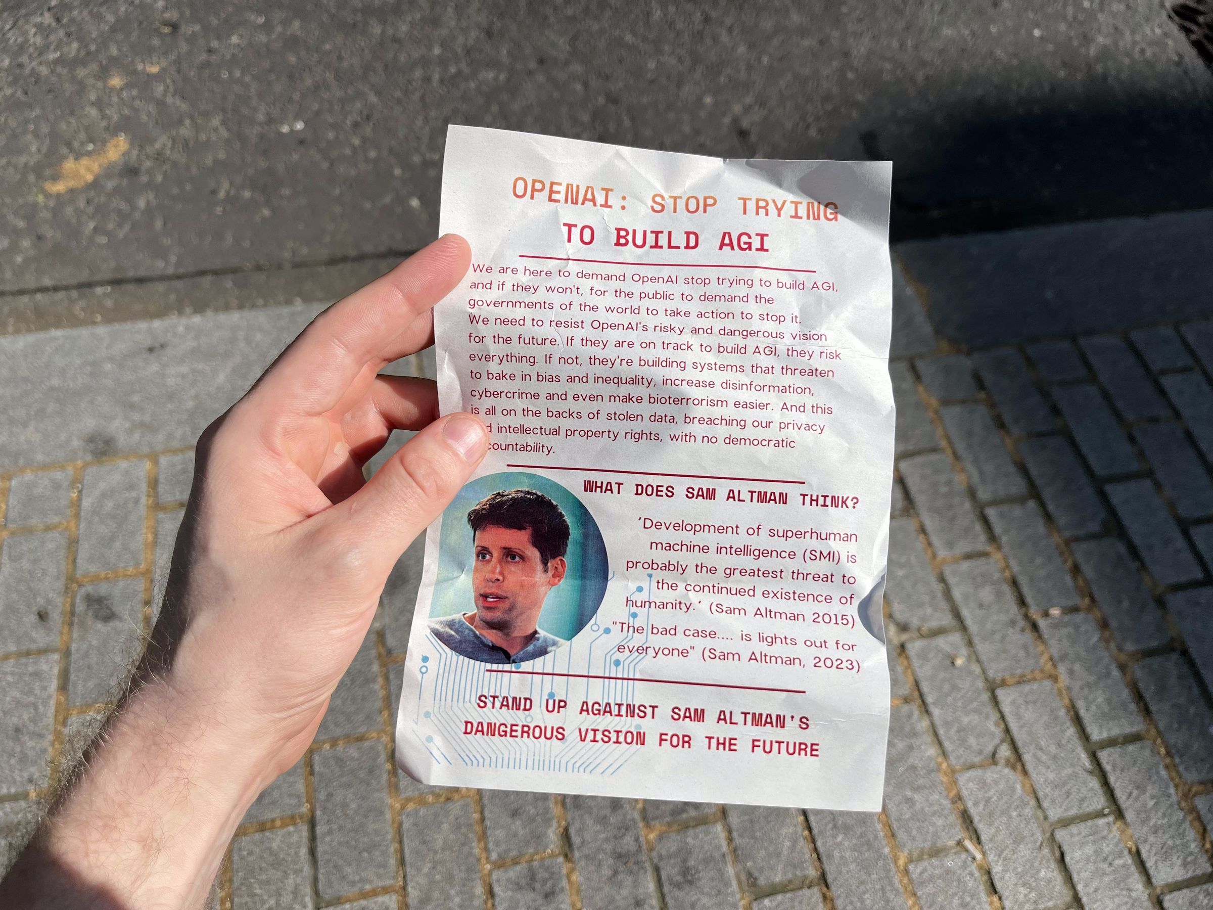 A photograph of a hand holding a leaflet with the title “OpenAI: stop trying to build AGI.” 