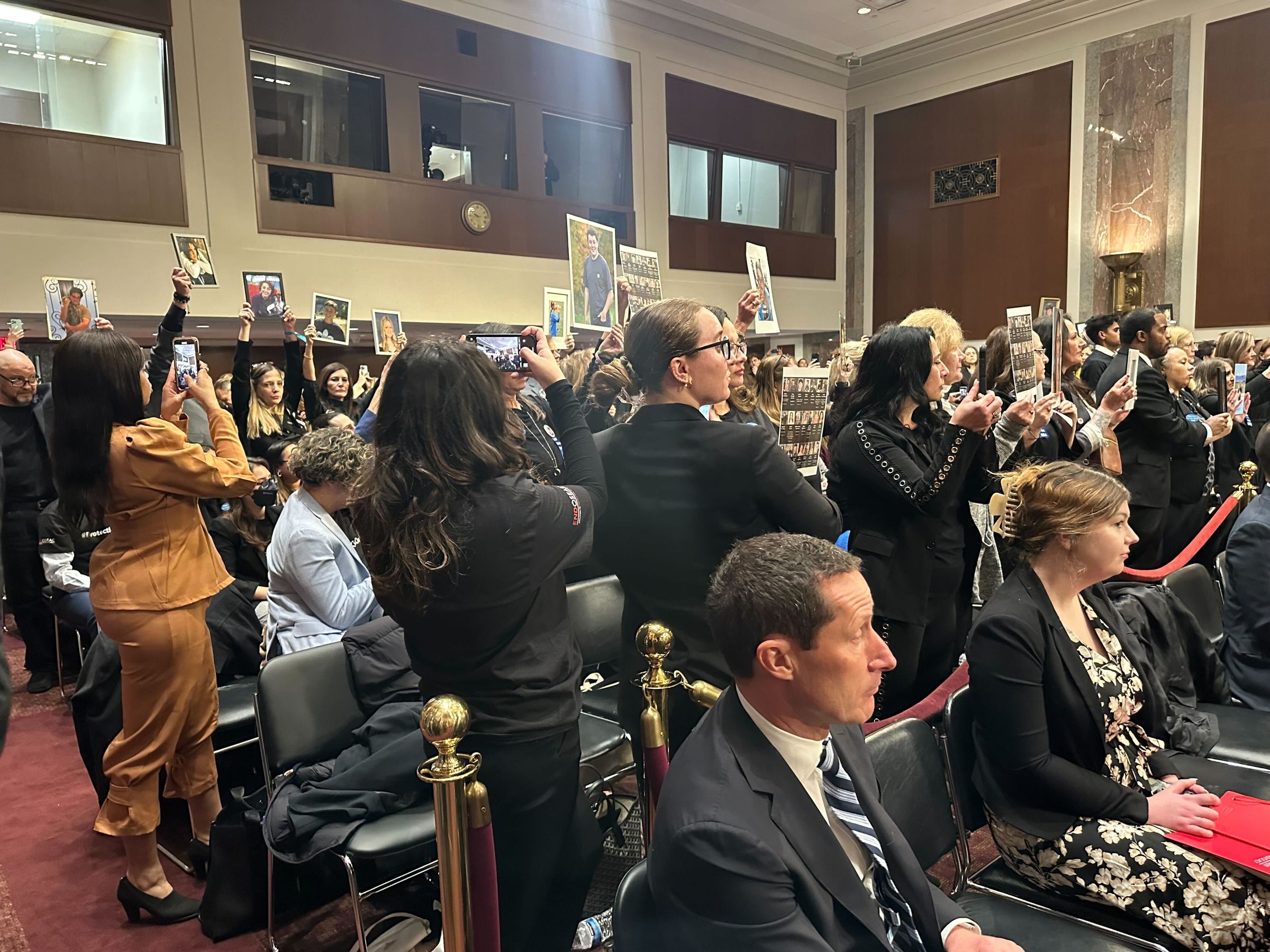 Advocates in hearing room awaiting tech CEO testimony stand with photos of teens and kids.