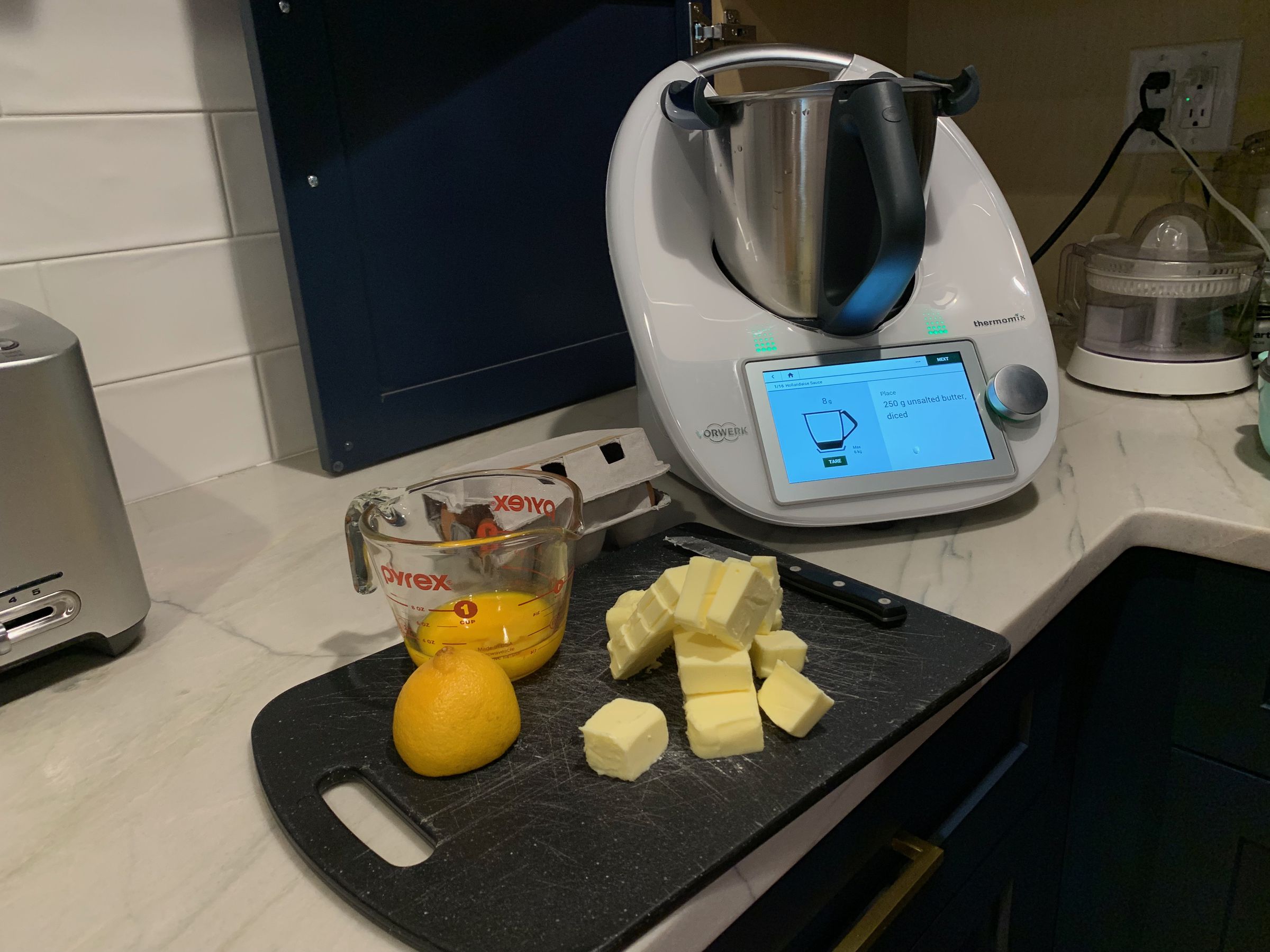 A counter with a measuring cup, some cut-up cheese, next to a Thermomix.