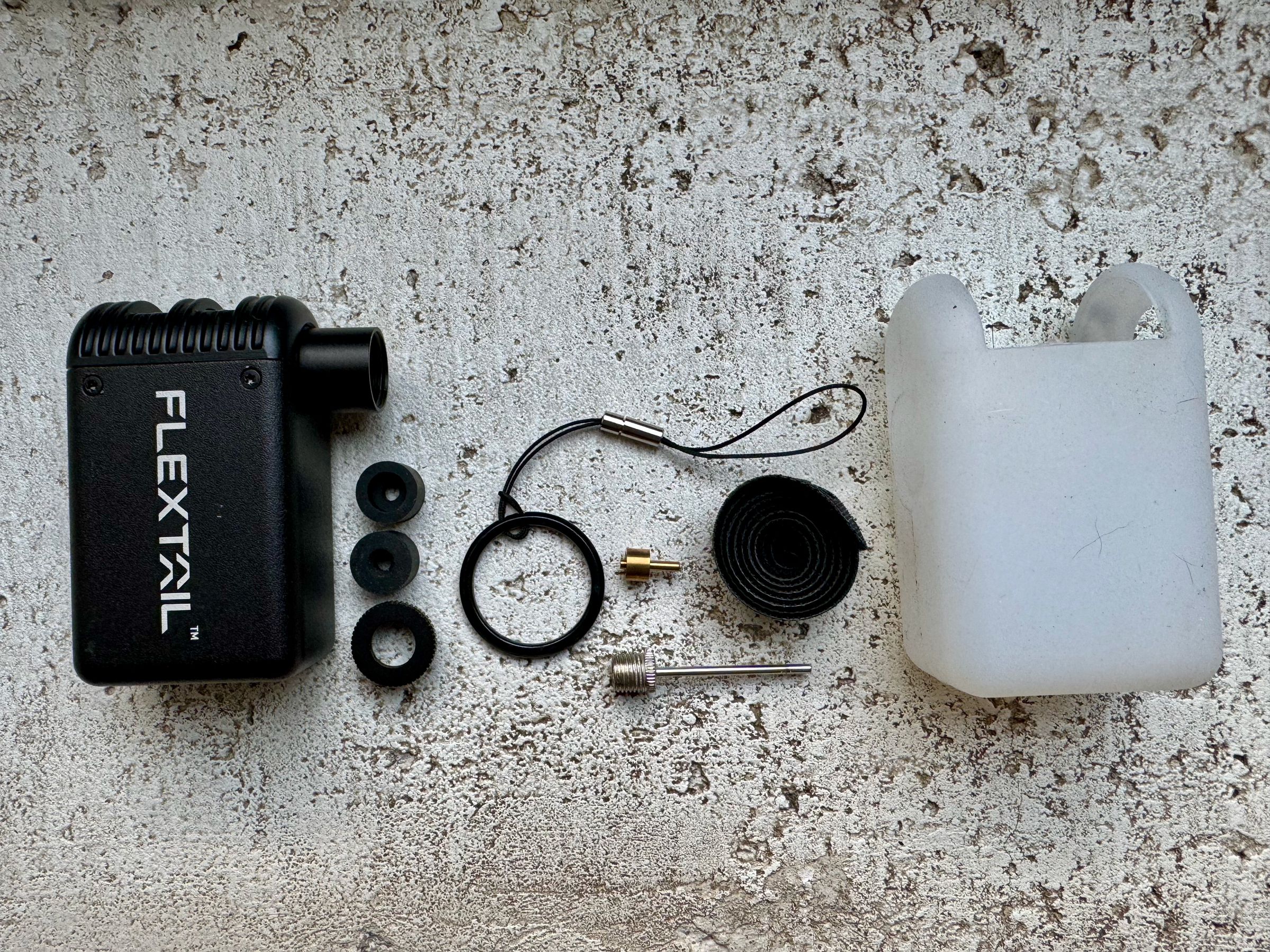 <em>From left to right: the Tiny Bike Pump, two rubber adapter valves and the enclosure that screws the adapters onto the pump nozzle, a carrying ring with strap, pin for Schrader valve adapter, ball pump nozzle, bike strap, and silicon sleeve. Not pictured is the USB-A to USB-C cable included in the box.</em>