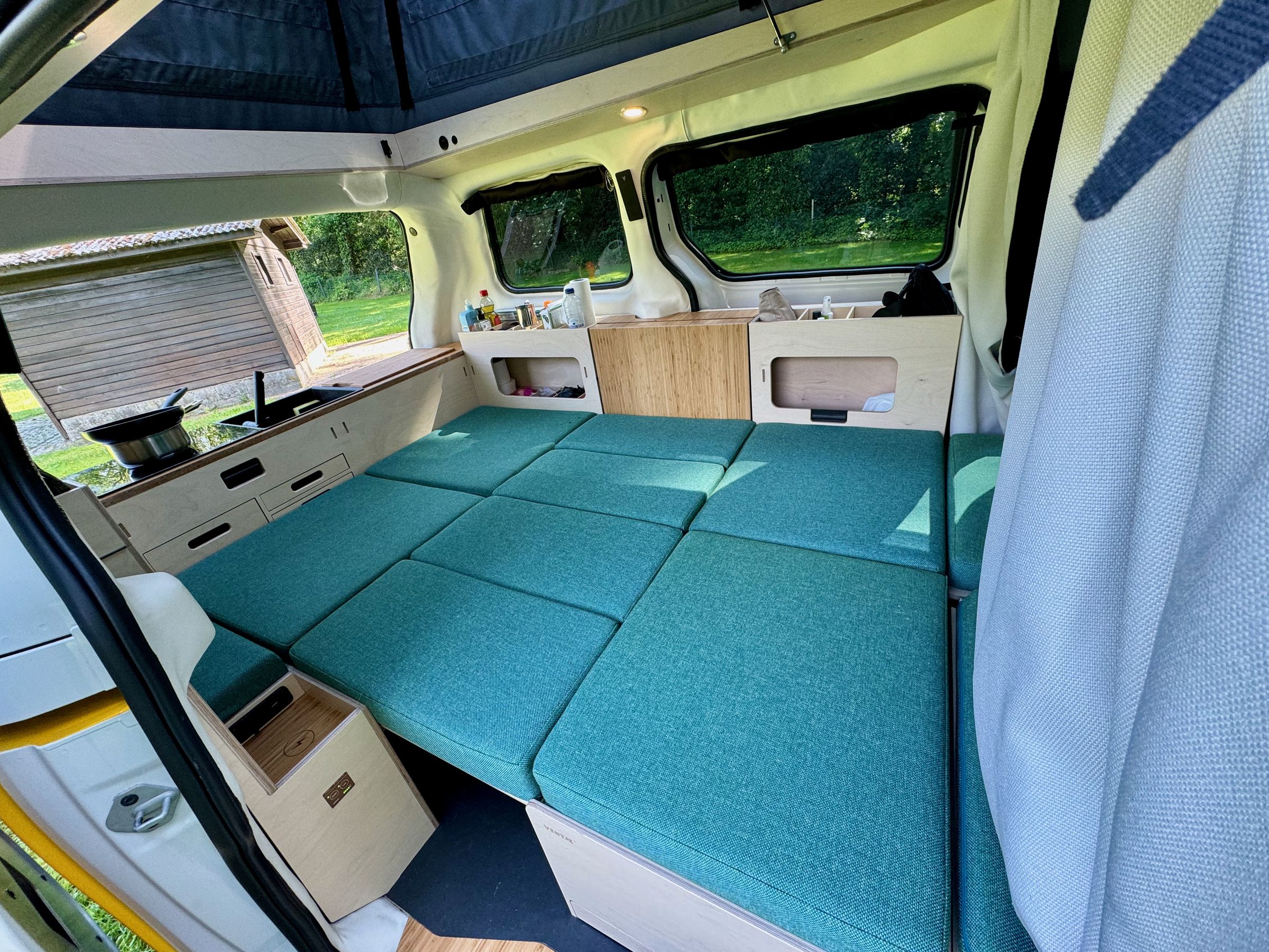 <em>Bed fully made from outside the van. All goes (mostly) dark when the magnetic window coverings are closed and the curtain is drawn between the sleeping area and driver and passenger chairs.</em>