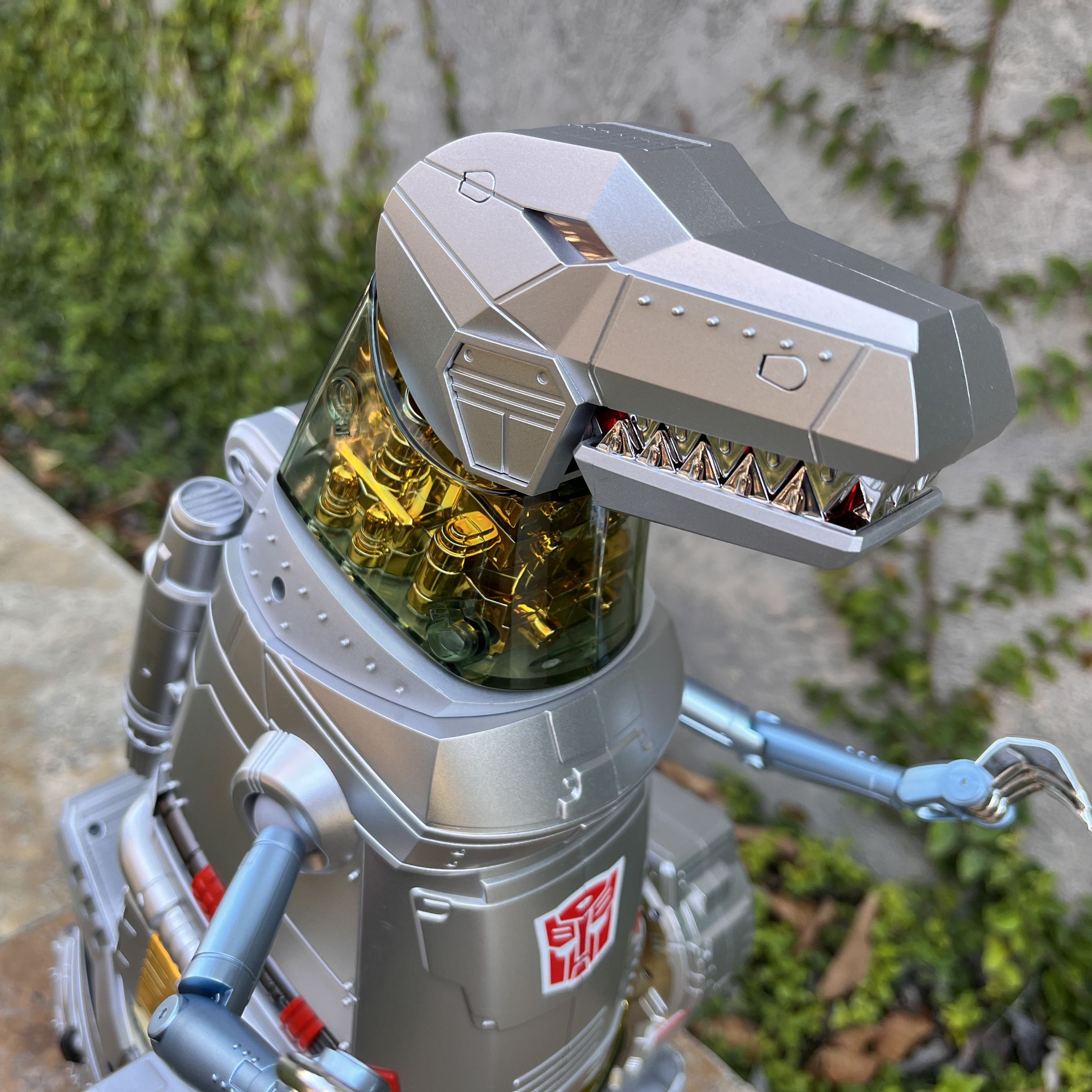 A silver robot T-Rex’s head with a see-through golden neck and tiny silver arms with three posable claws each.