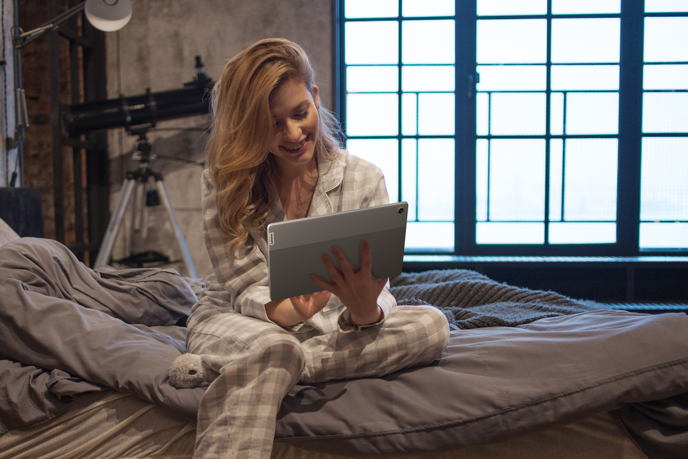 A user sits in pajamas on a bed with a bright window in the background, using the Lenovo Chromebook Duet 5 in tablet mode.