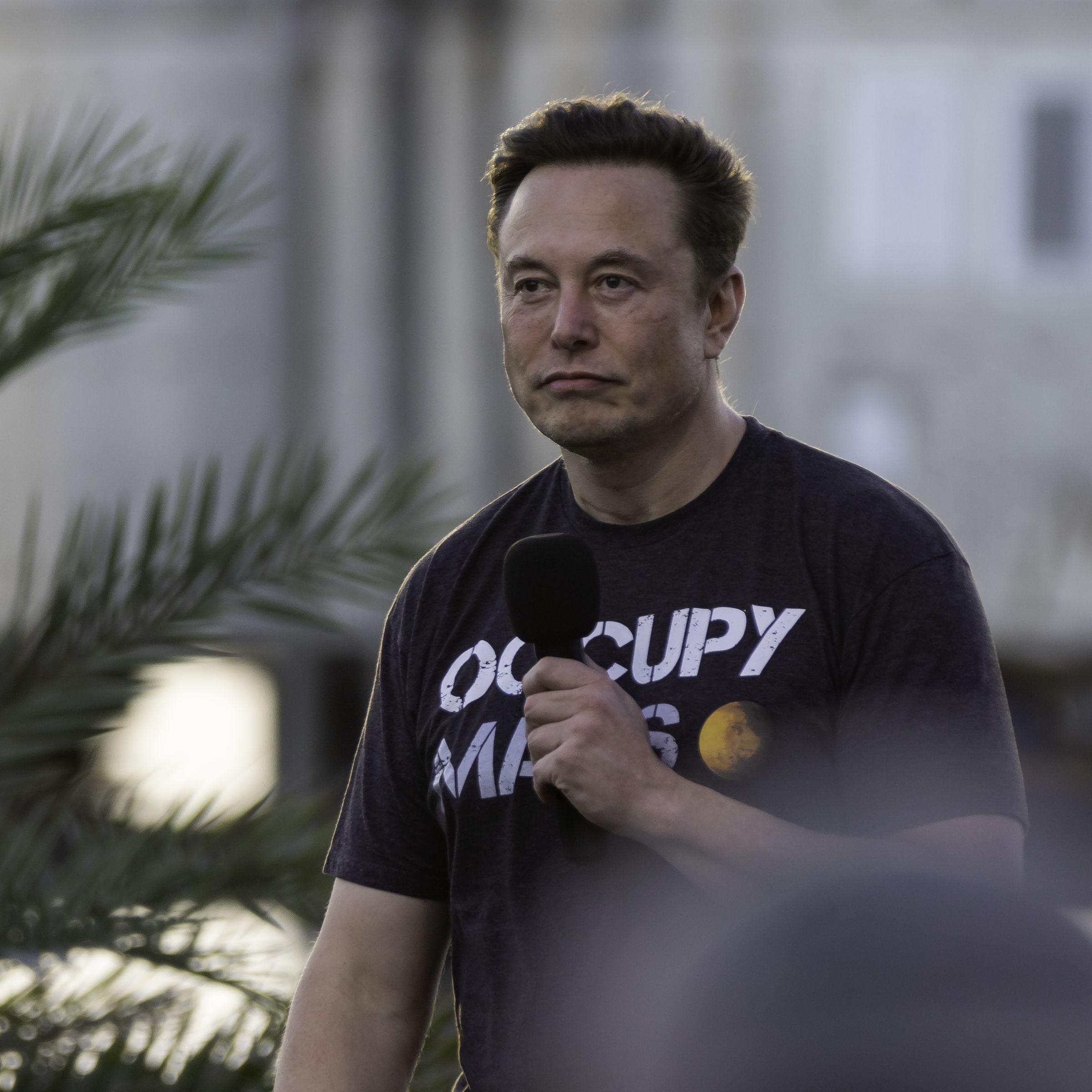 Elon Musk holds a microphone to his face while wearing a T-shirt that says “Occupy Mars.”
