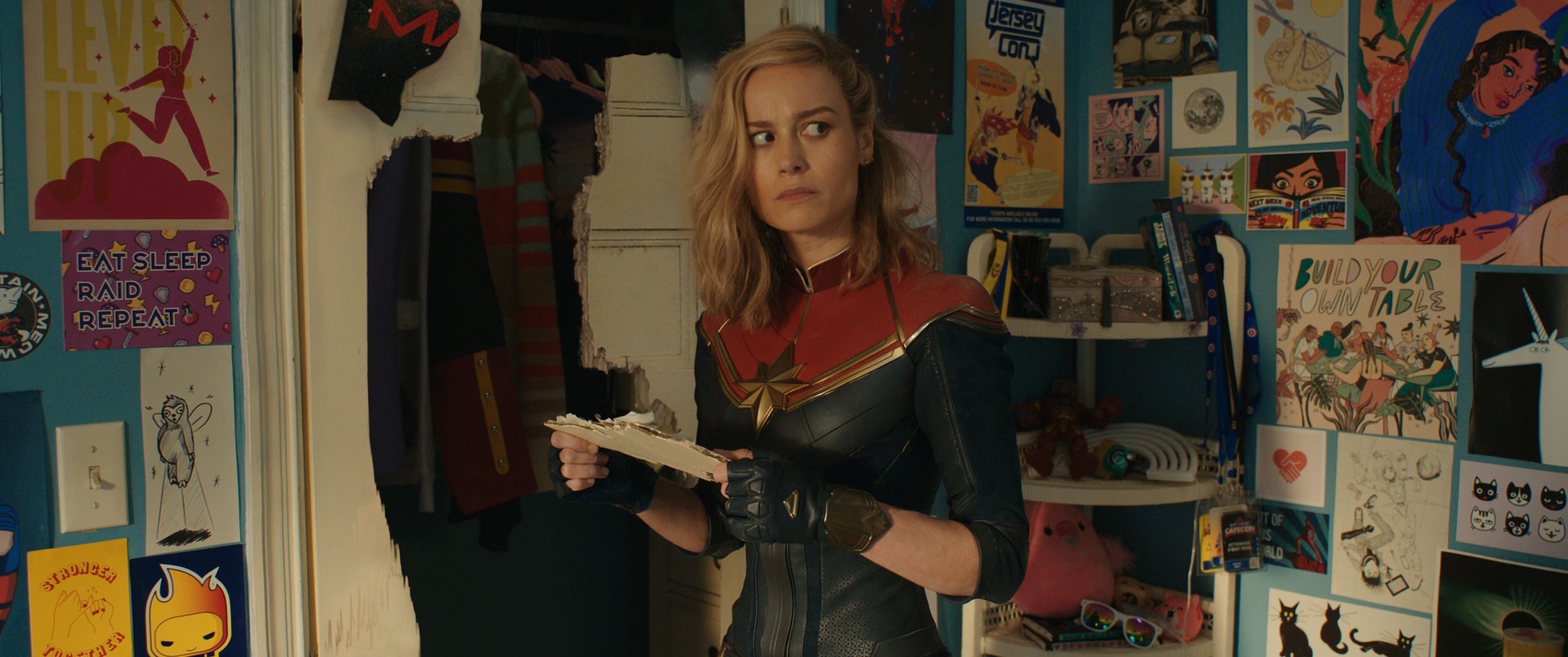 A woman in a form-fitting superhero costume standing in a bedroom whose walls are plastered with drawings of her.