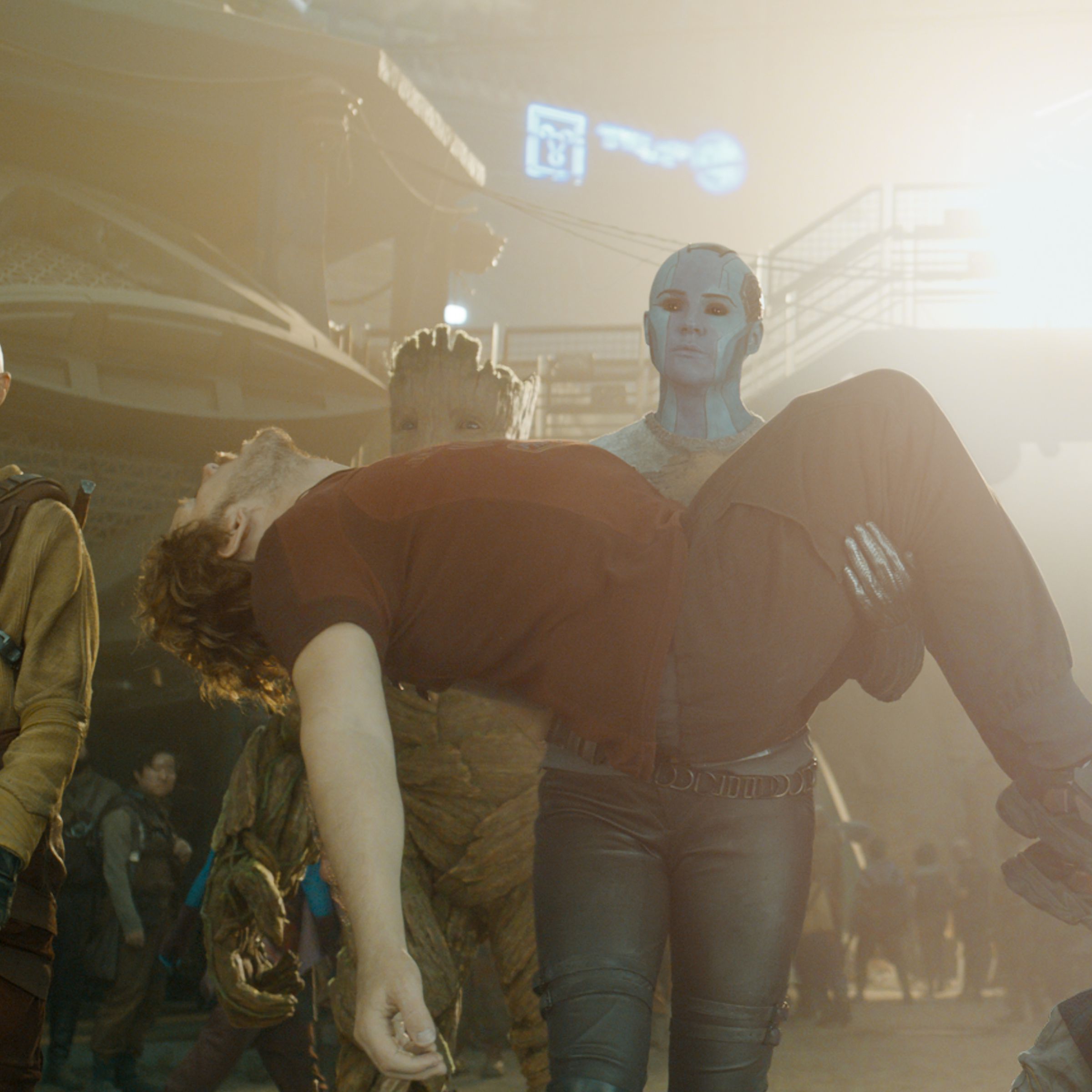 An image of the Guardians from Guardians of the Galaxy. Nebula, a blue cyborg woman, stands in the foreground bridal carrying an unconscious Peter Quill, a large muscular white man.