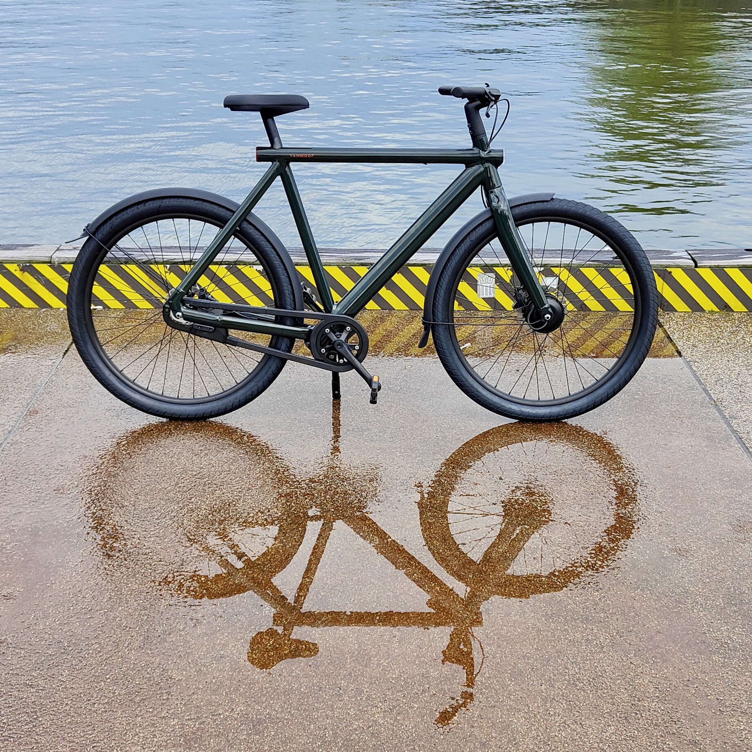 A dark green VanMoof S4 e-bike stands on a wet concrete platform surrounded by water and yellow and black warning paint around the platform’s permitter. The bicycle is reflected in the wet concrete.