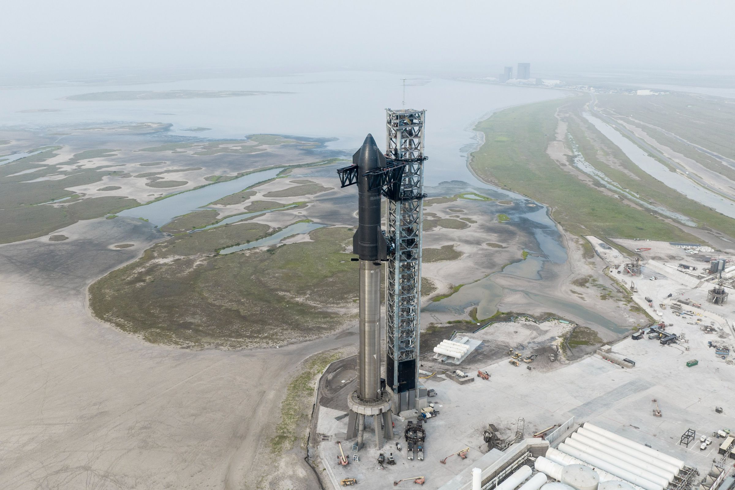 Zoomed-out landscape of SpaceX’s Starbase in Boca Chica, Texas, showing a water landscape in the back and the rocket front and center.