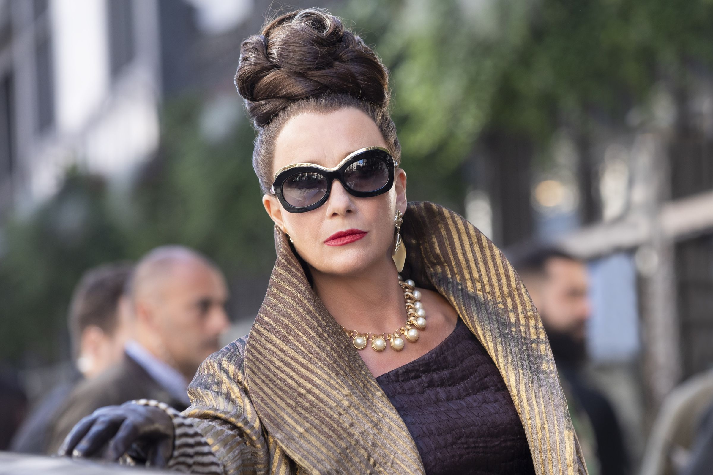 Emma Thompson as the Baroness. Her hair is up in an intricate hairstyle, she is wearing large sunglasses, gold earrings, a pearl necklace and a large gold coat.