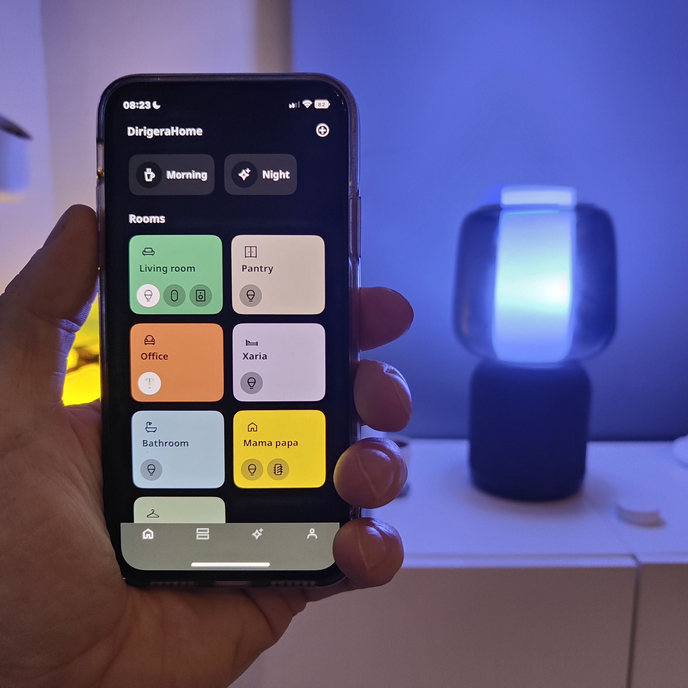 An iPhone running the new Ikea Home smart app held in front of the Symfonisk desk lamp, Dirigera hub, and Sound Remote.