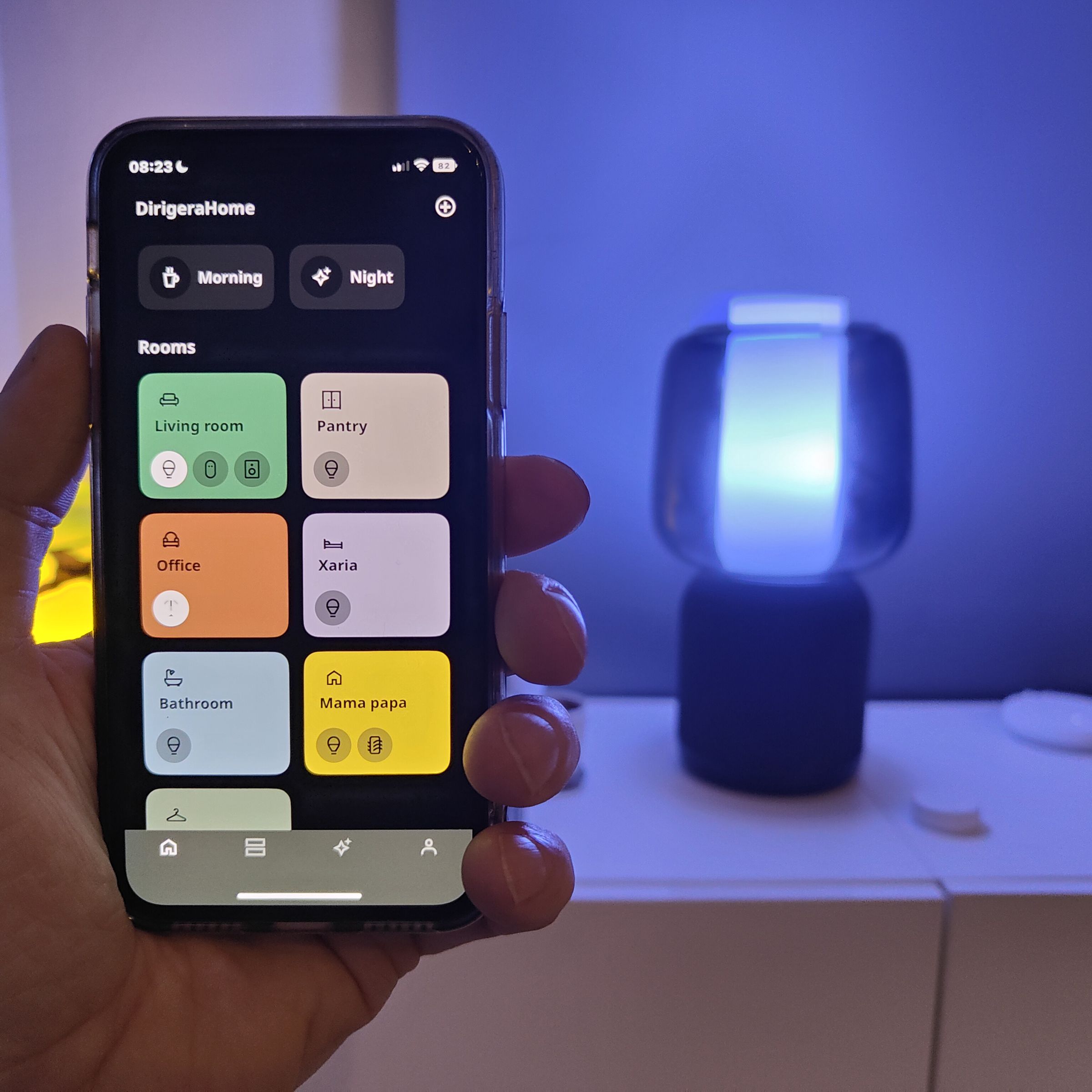 An iPhone running the new Ikea Home smart app held in front of the Symfonisk desk lamp, Dirigera hub, and Sound Remote.