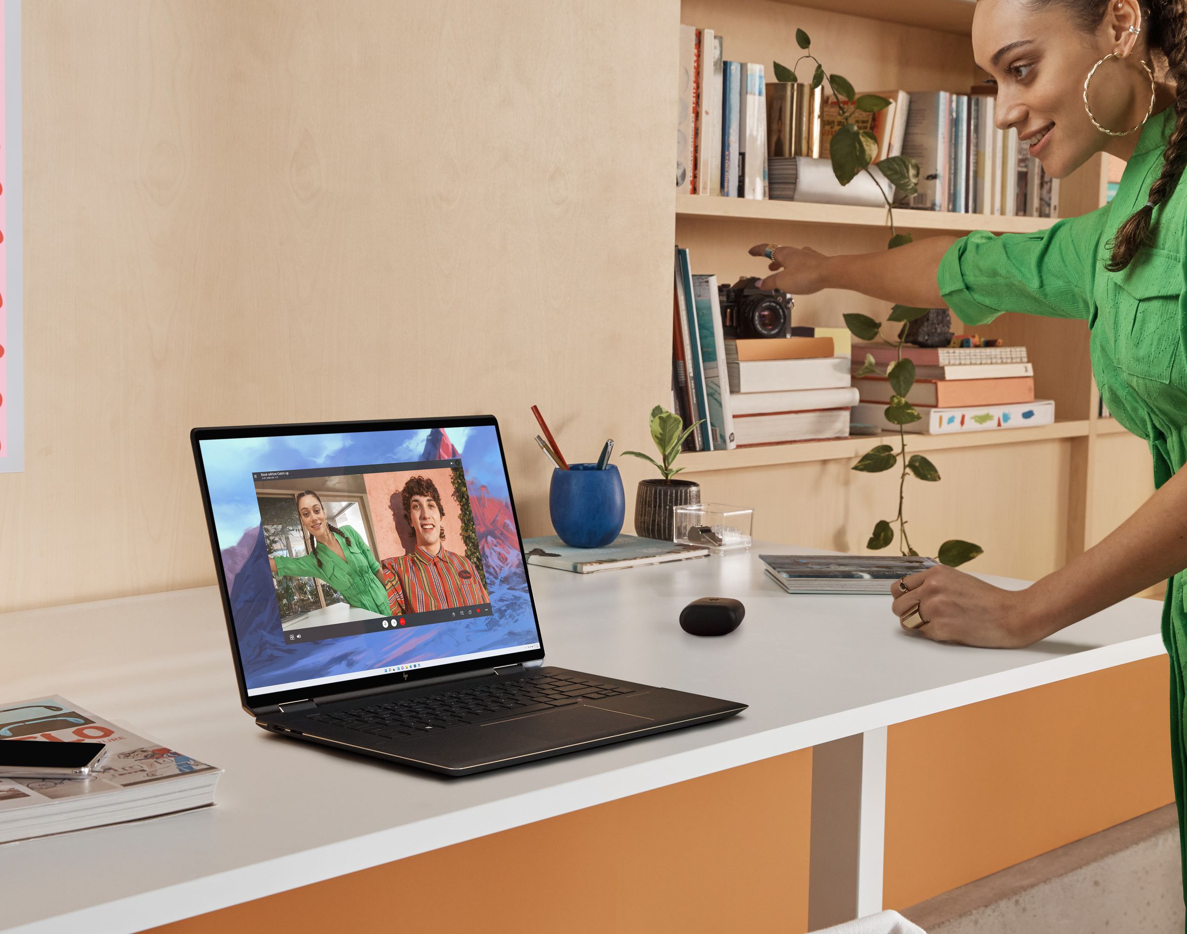 A user participates in a video call on the HP Spectre x360 16, pointing to the right. The laptop sits on a white desk. To its left are a book and a smartphone. To its right are a small potted plant, an earbuds case, a pencil case, and a full bookcase.