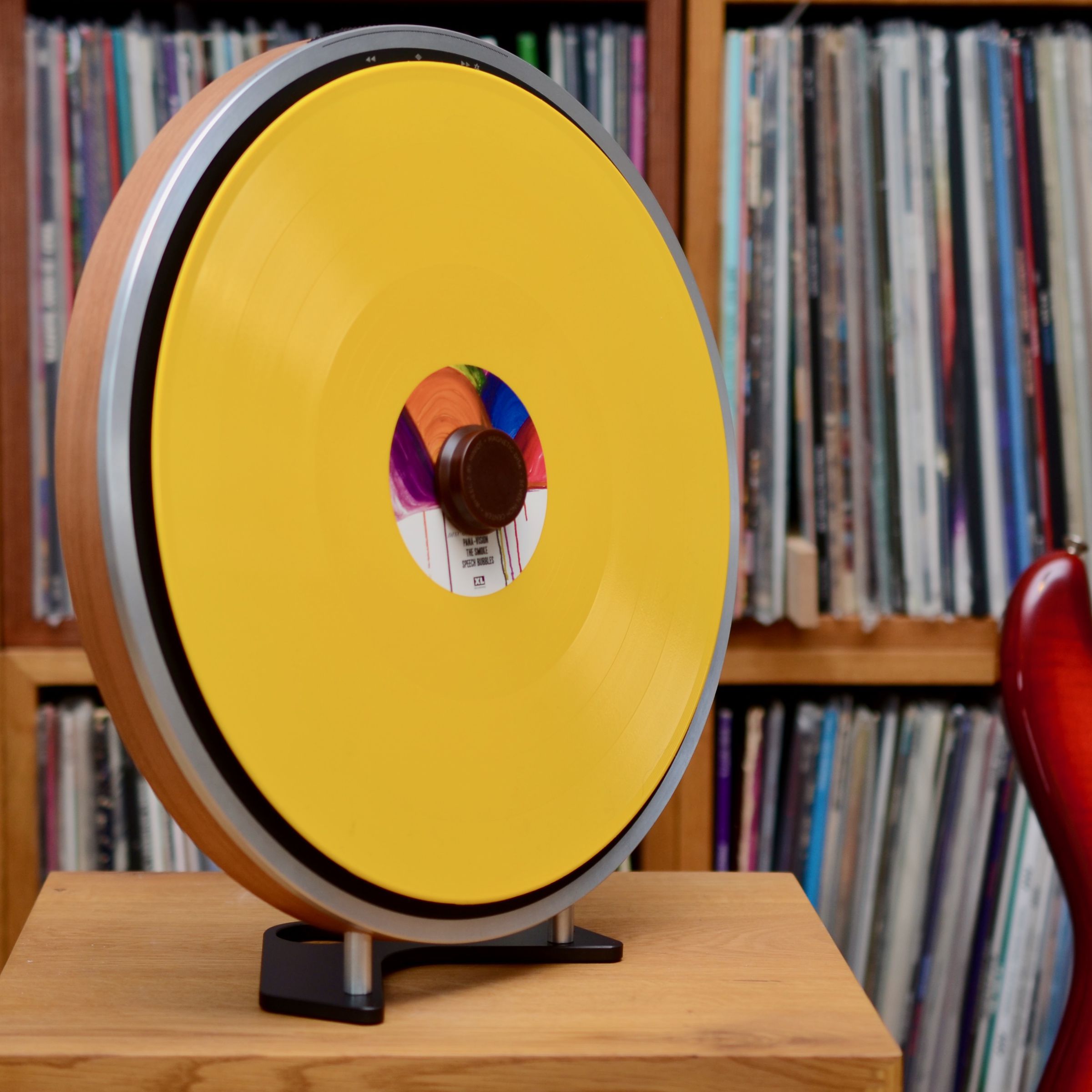 The Miniot Wheel 2 turntable stands upright with a bright yellow record installed. Behind it is a wall of albums inserted into boxes with a red guitar leaning against it.