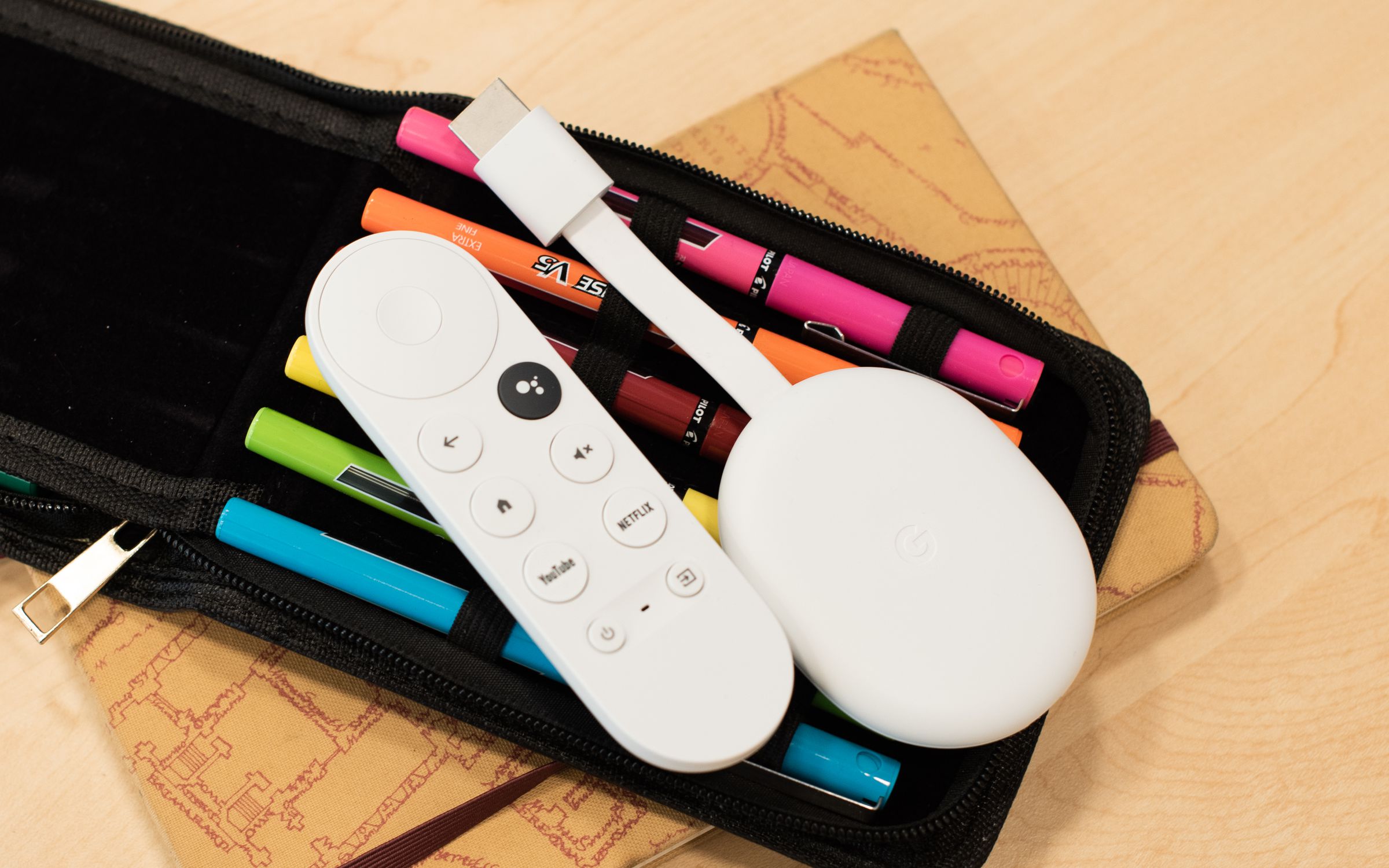 Image of a Chromecast with Google TV sitting on a bag of pens. It’s a white, oval-shaped device, with an HDMI cable sticking out from the front.