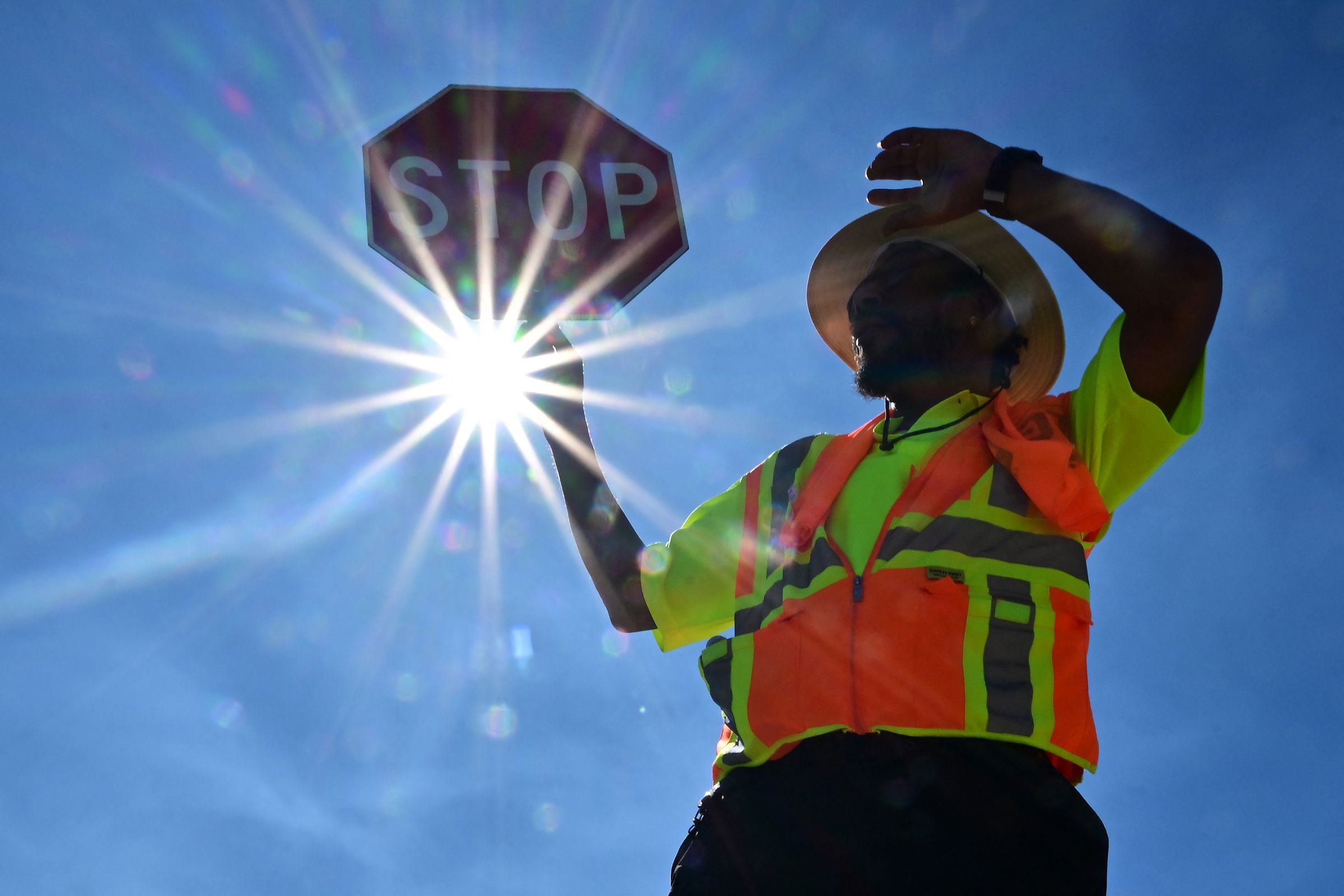 A person wearing an orange work vest holds up a stop sign. The sign shines brightly behind them.