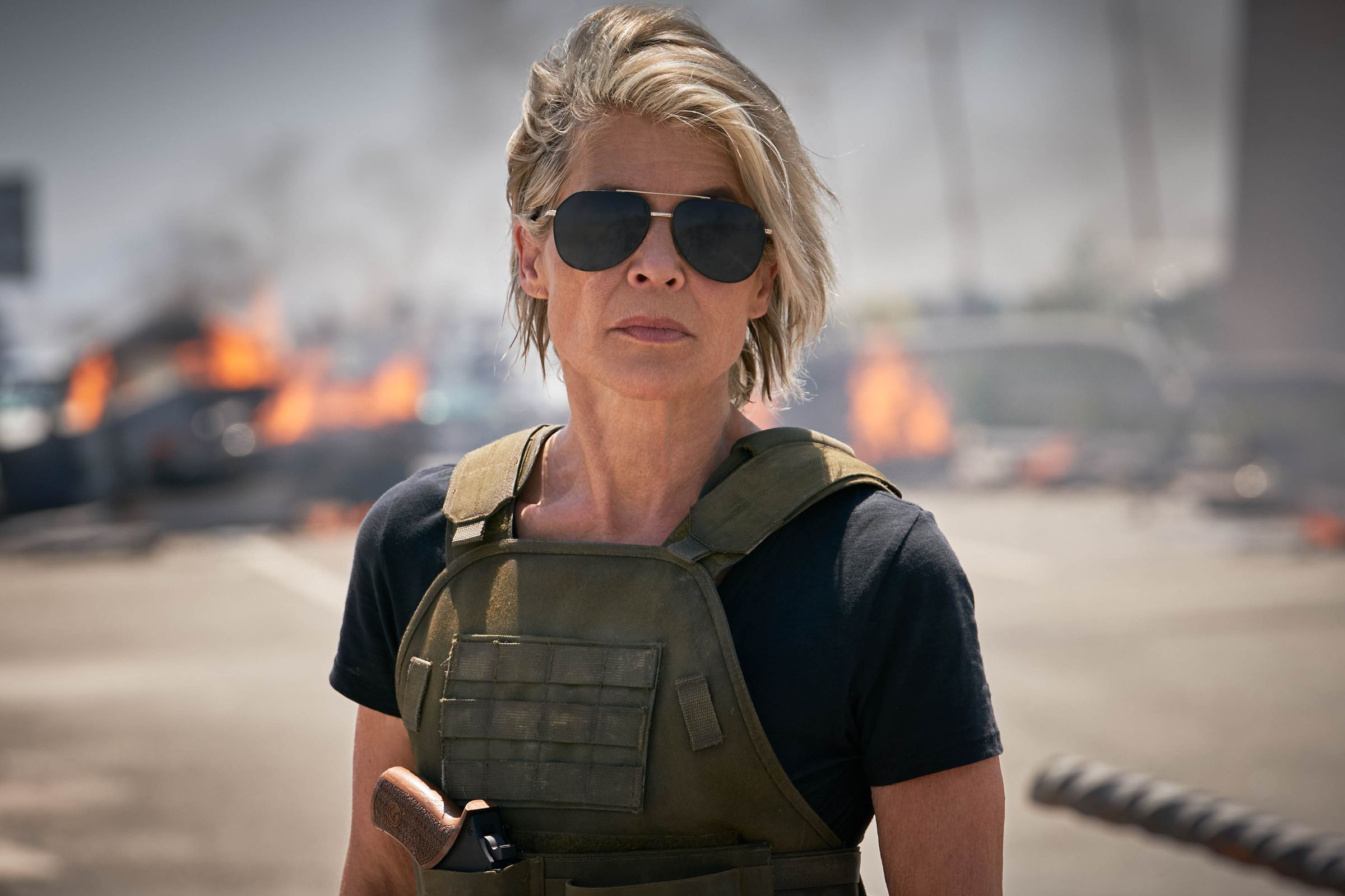 Linda Hamilton, wearing a combat vest and aviator classes, looking non-plussed. 