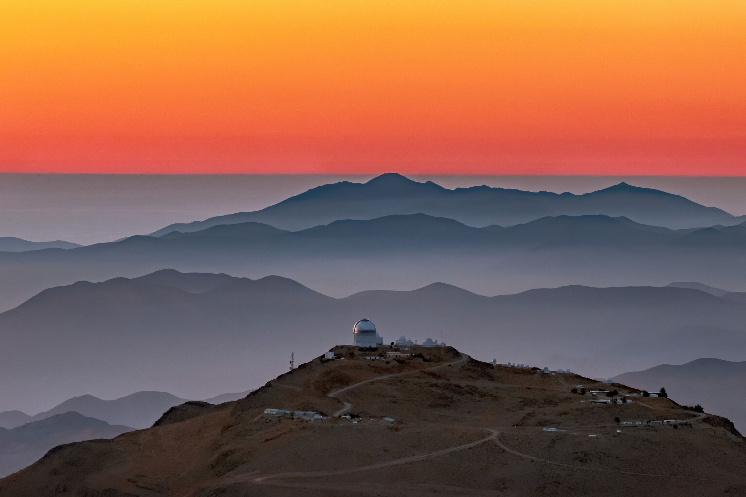 The domes of a cluster of satellites sits on a mountain ridge in the foreground. Mountain ridges rise in the background, transitioning into a orange sunset sky.