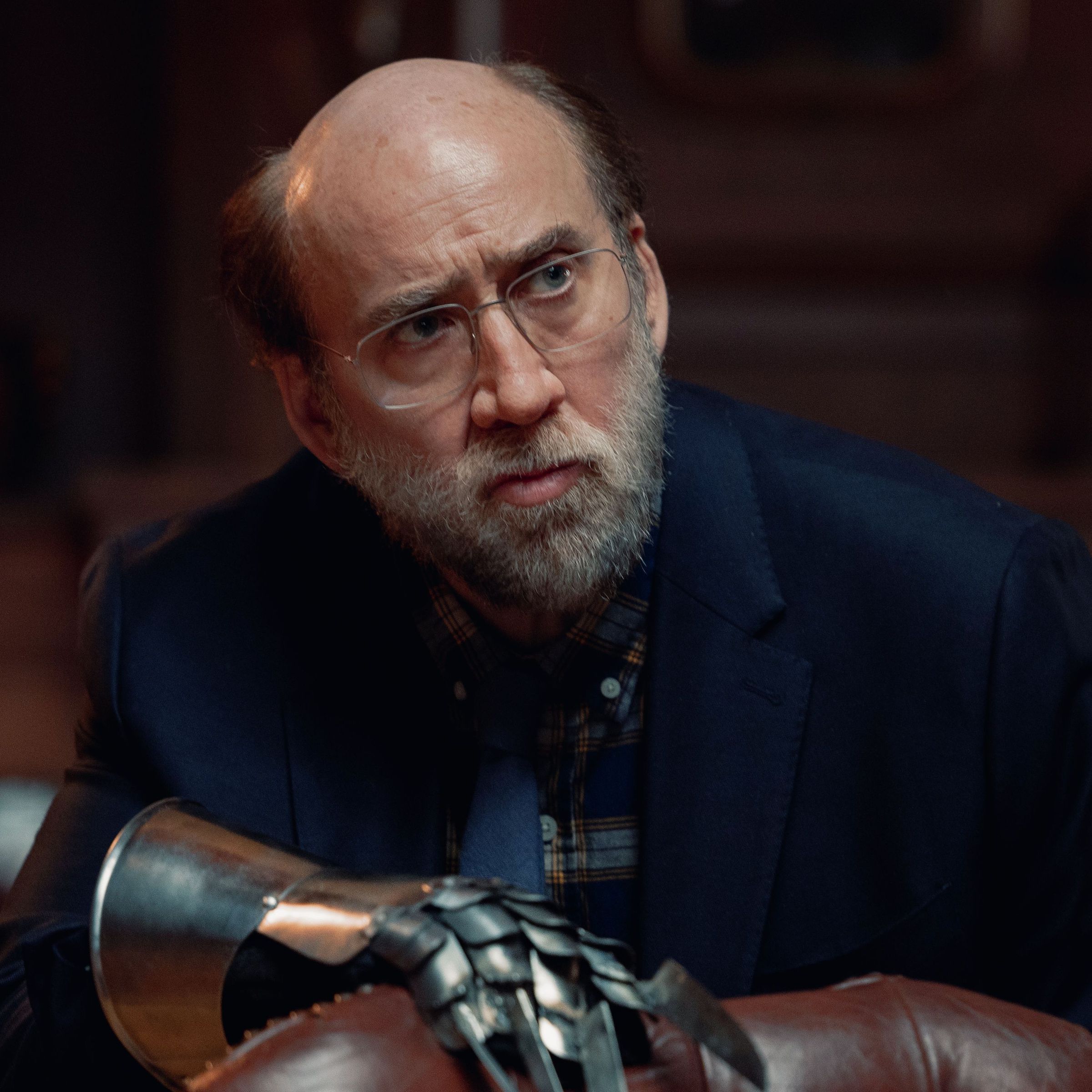 A balding middle-aged man wearing glasses, a blue suit, and an armored glove on one hand as he leans over to rest his arm on the back of a leather chair.