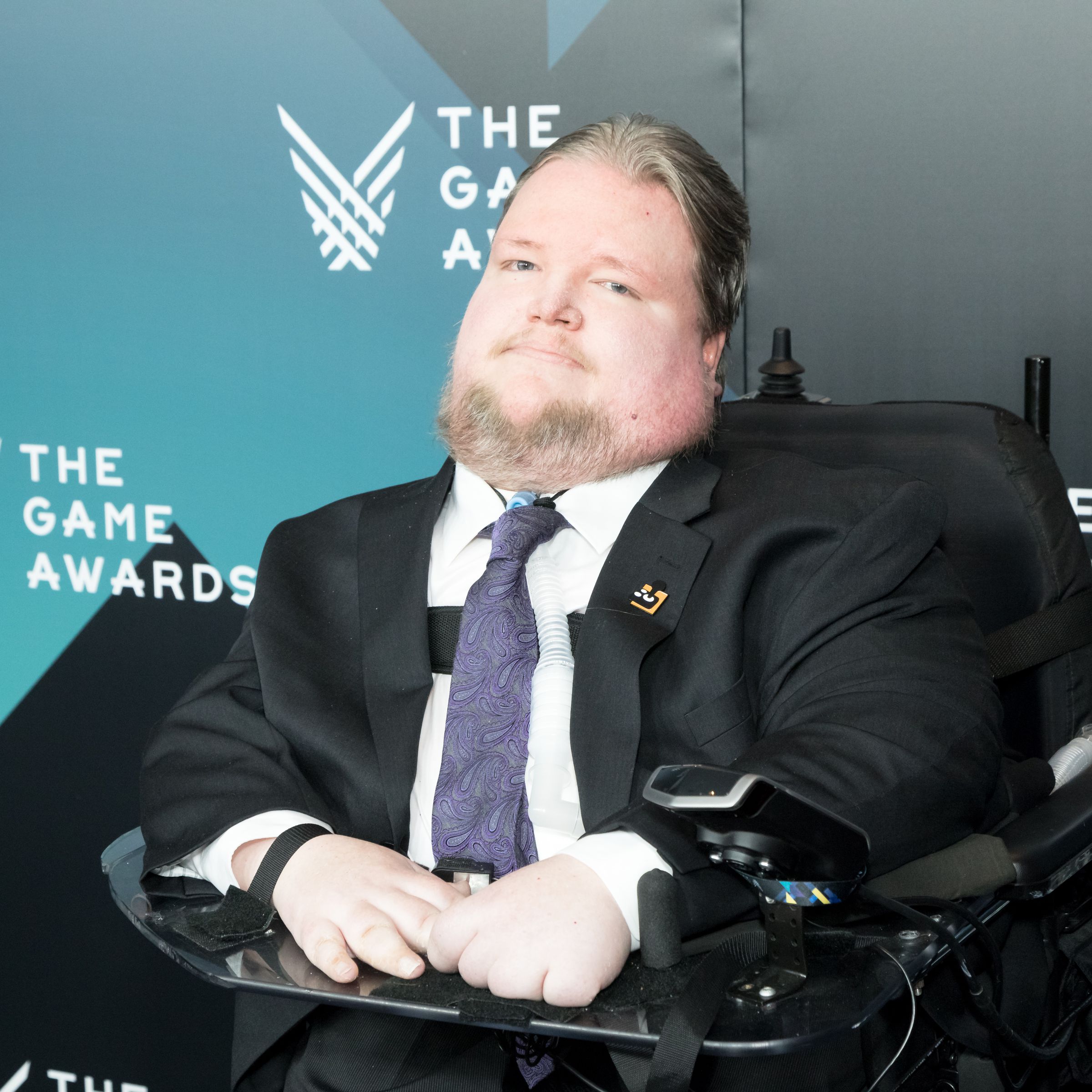 Steven Spohn wearing a suit, sitting in his wheelchair in front of a backdrop with “The Game Awards” on it. 