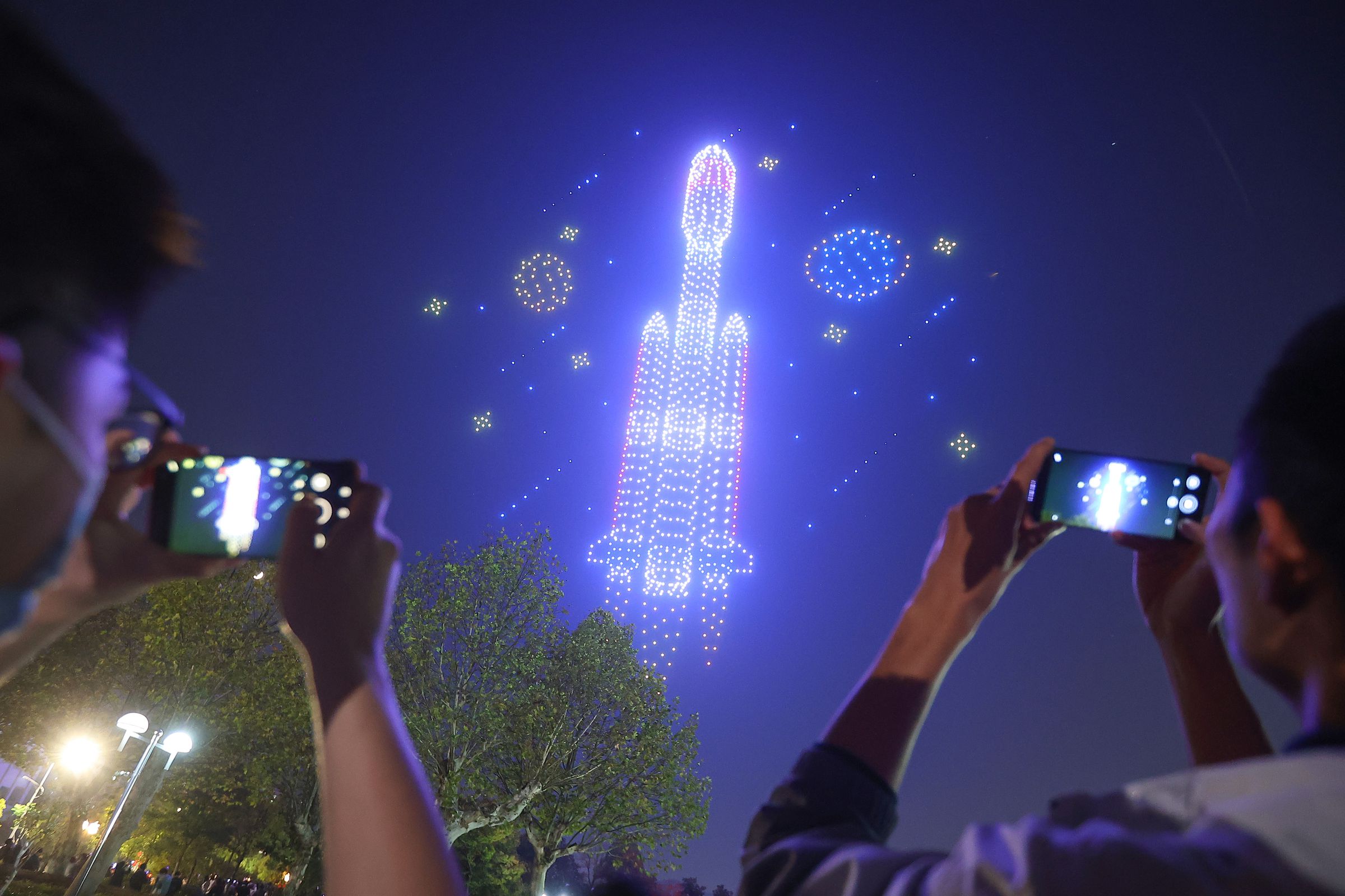 2,022 drones performing in a light show to celebrate the 70th anniversary of a local university in Nanjing, China. The drones have formed a rocket, and bystanders are taking pictures on their mobile devices.