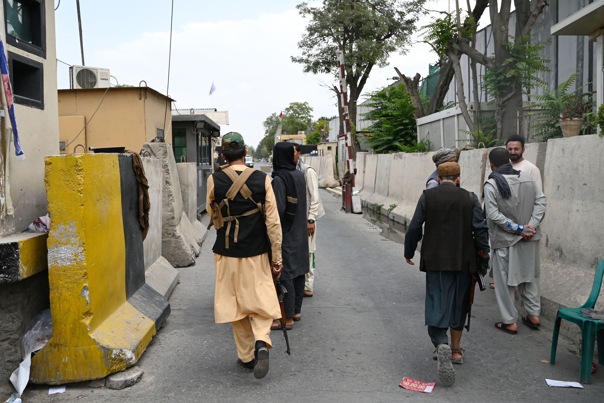 Four men facing away from the camera carry military rifles along an empty street, flanked by concrete barriers.
