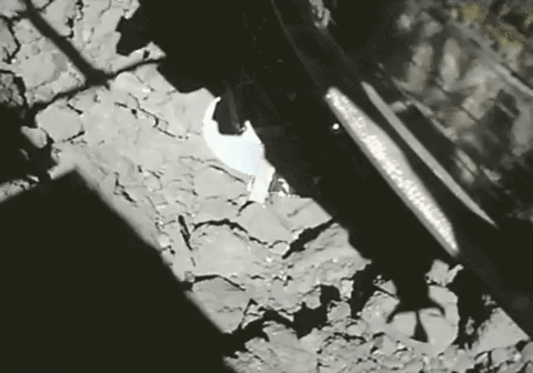 A rectangular shadow approaches a rocky surface, it appears to touch the surface sending dust and rocks flying upwards towards the camera.