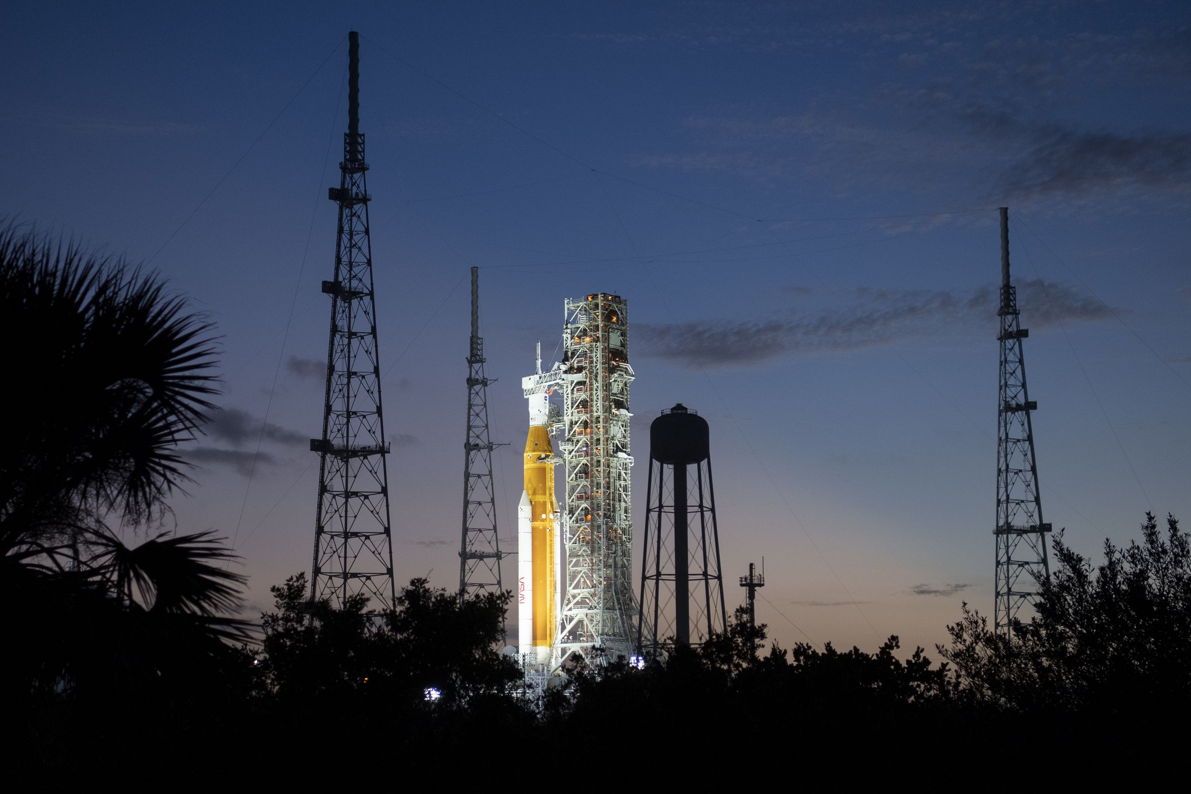 An illuminated rocket sits on a launchpad surrounded by equipment, silhouetted against a dusk sky.
