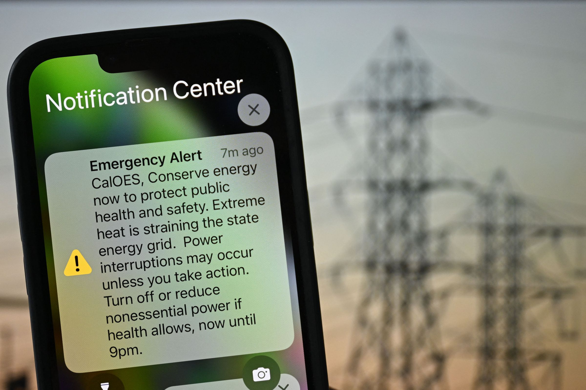 iPhone alert screen showing an emergency alert to conserve energy in California, against the backdrop of power lines.