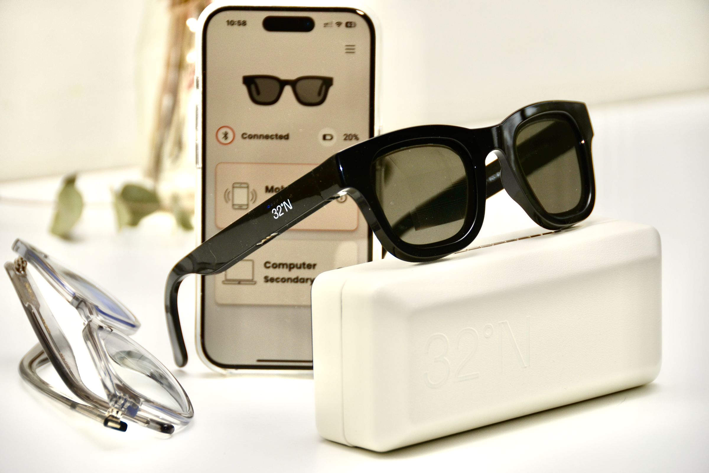 A pair of Muir sunglasses sit perched on top of their case in front of an iPhone open to the 32°N app showing 20 percent battery remaining on the sunglasses.