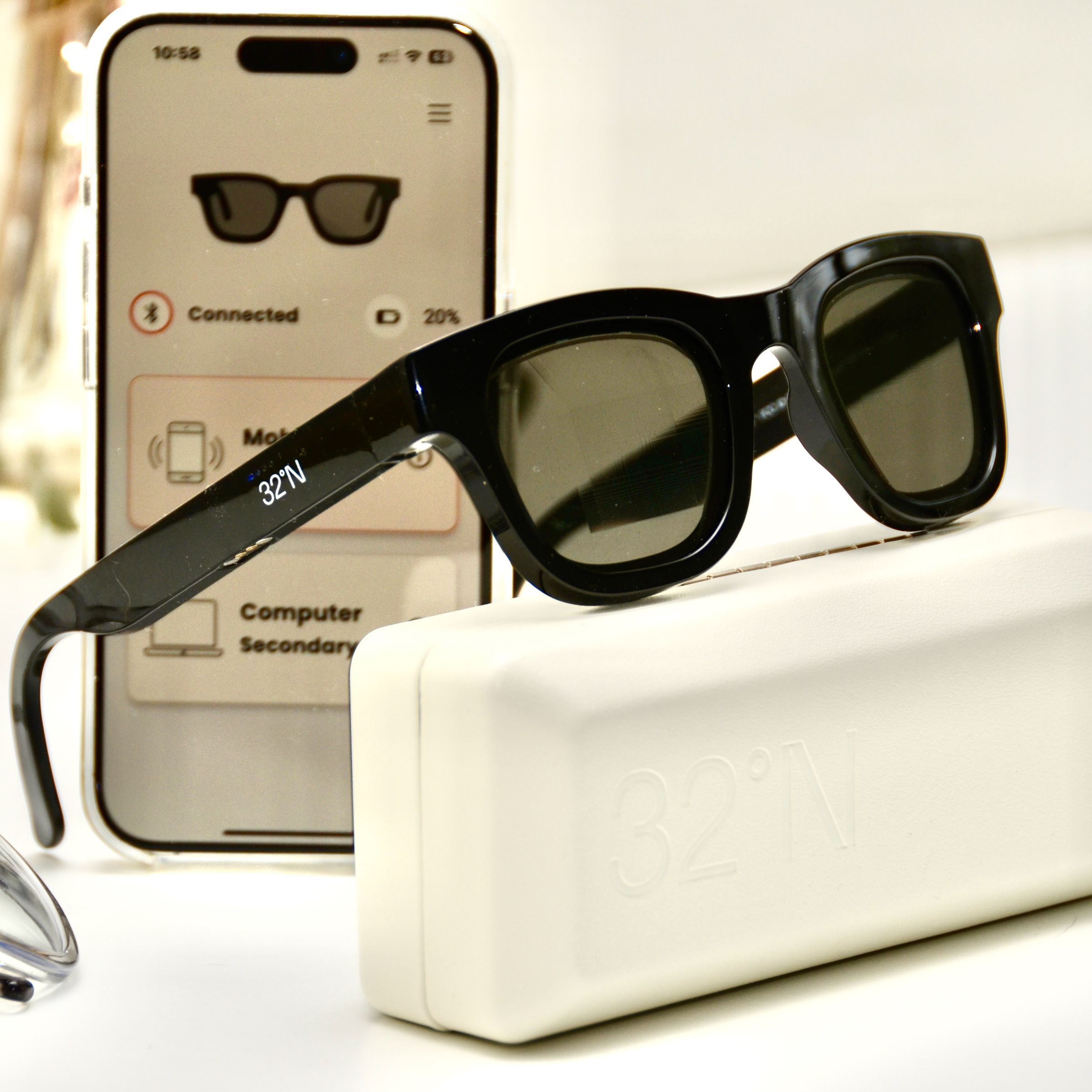 A pair of Muir sunglasses sit perched on top of their case in front of an iPhone open to the 32°N app showing 20 percent battery remaining on the sunglasses.