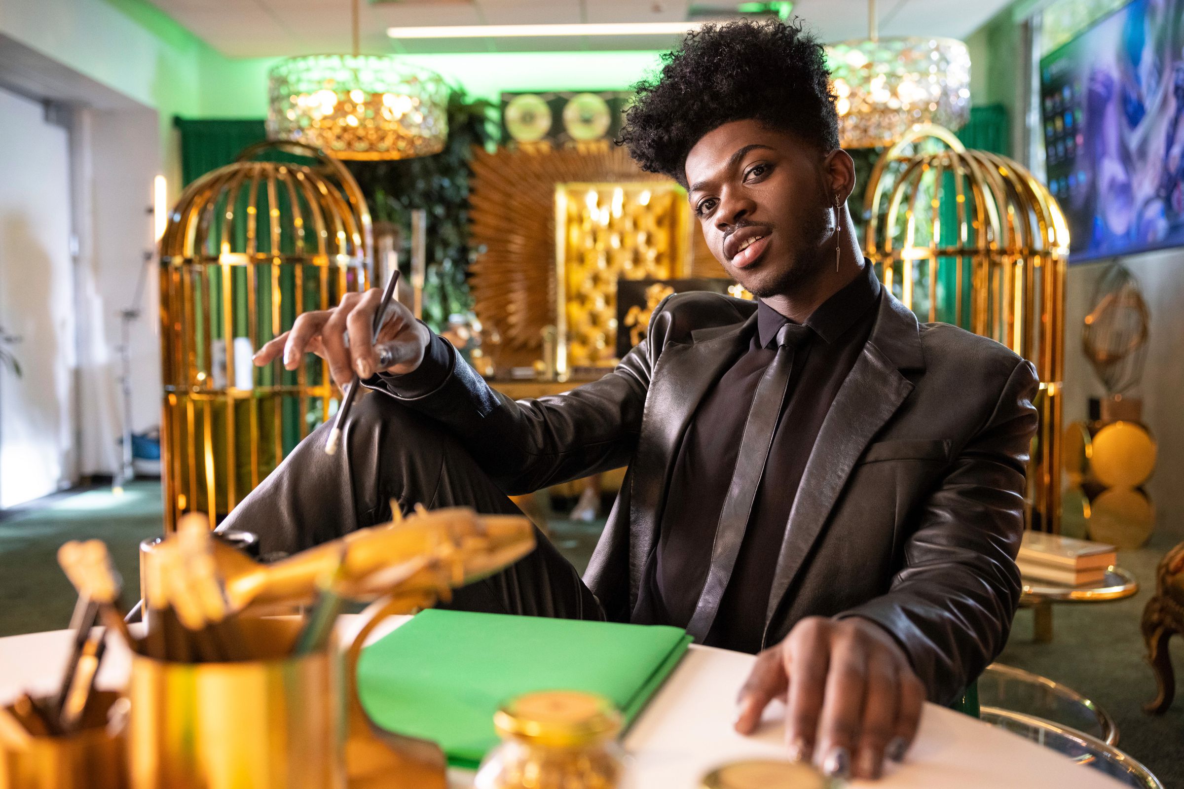 A photo of Lil Nas X in an all-black outfit, seated at a table in a room filled with gold accents.