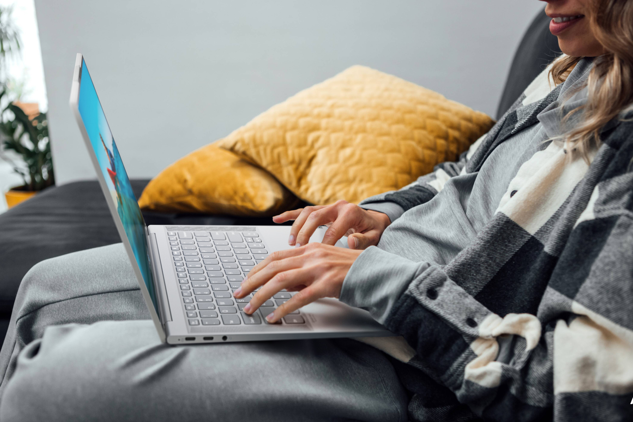 A user sits on a couch in sweatpants and a flannel, seen from the side, typing on an IdeaPad Slim 7 Carbon. 