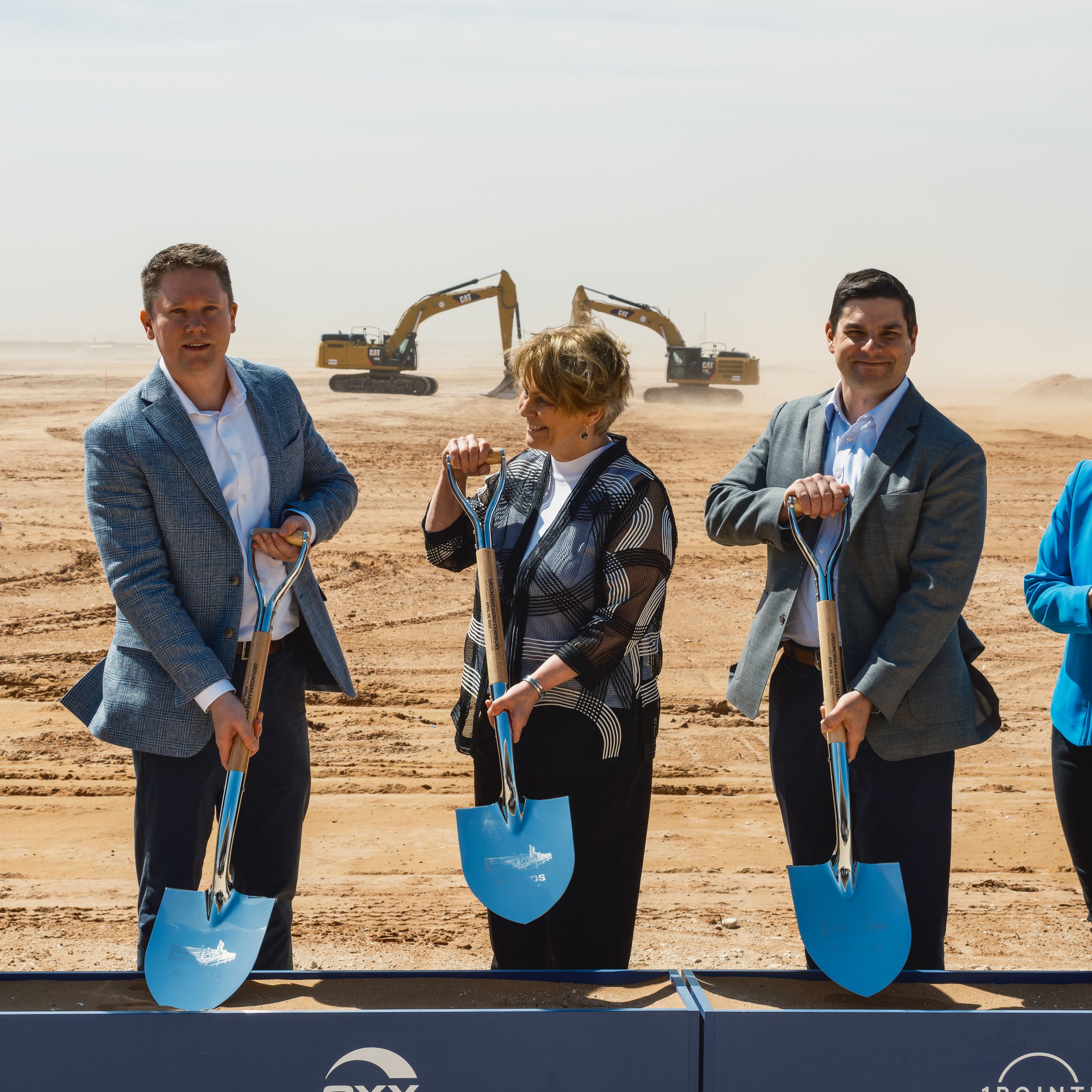 A row of people wearing suits hold up shovels at a construction site.