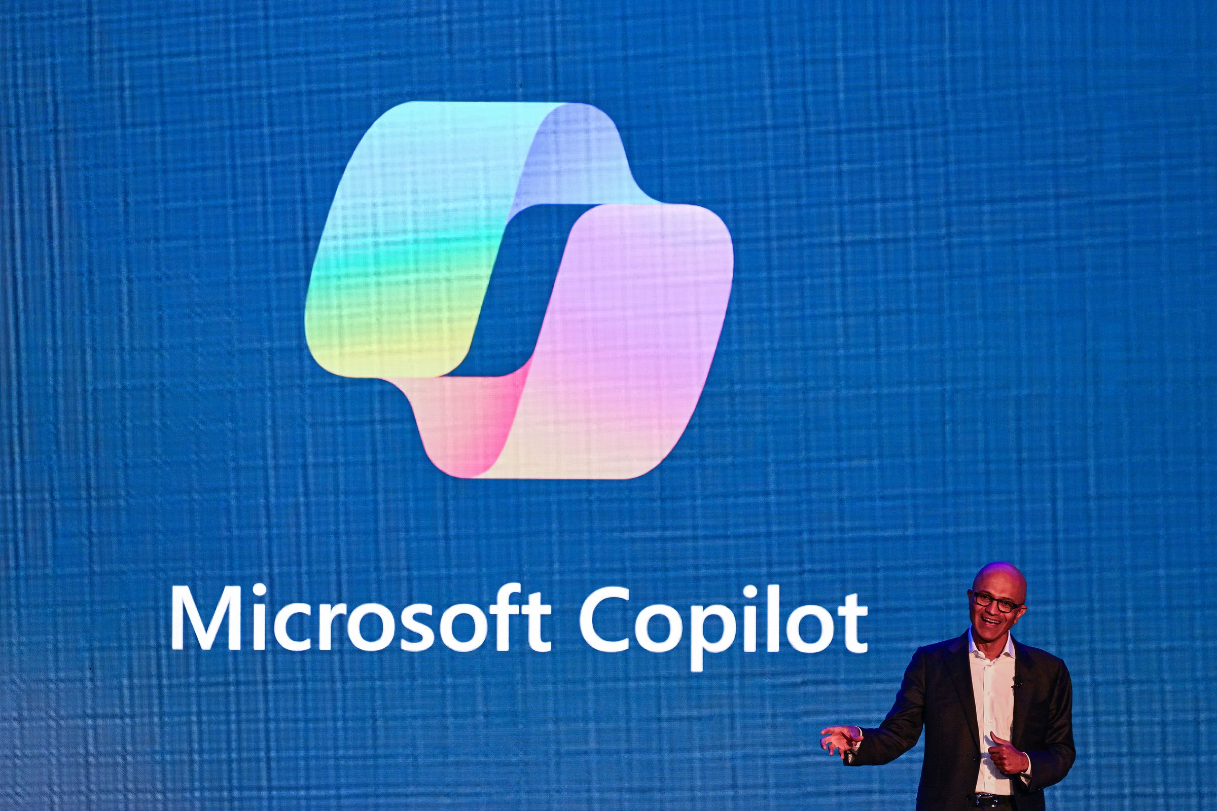 A man wearing a suit stands in front of a large screen that says “Microsoft Copilot”