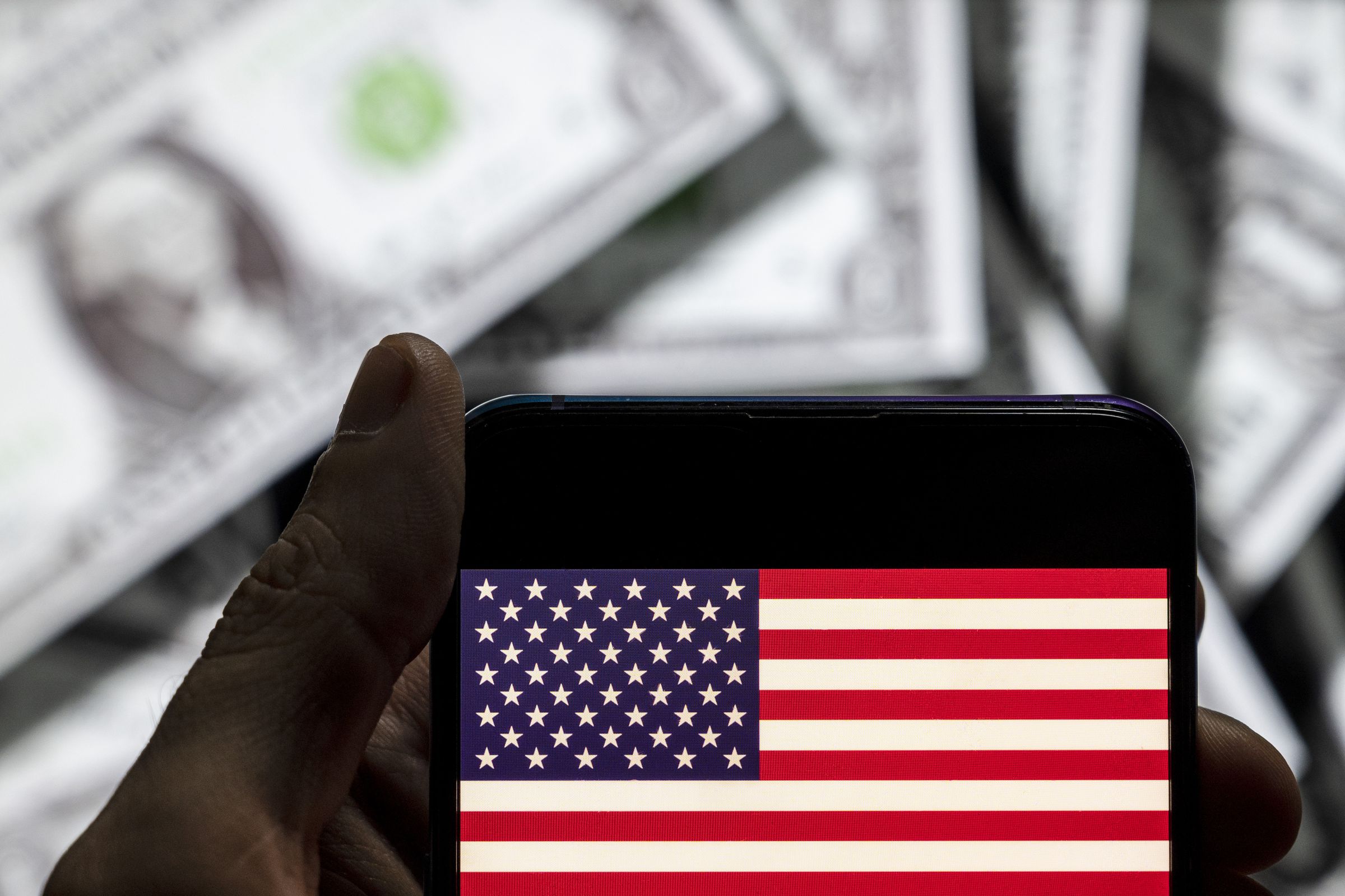 In this photo illustration the United States of America flag seen on a phone with dollar bills behind it.