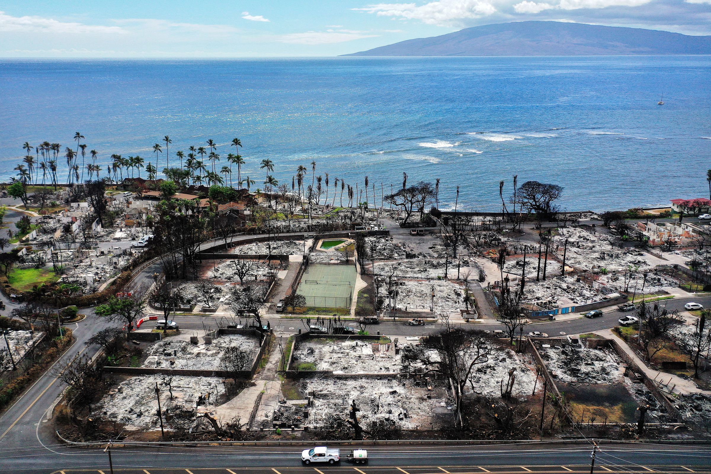 In an aerial view, a recovery vehicle drives past burned structures and cars in a neighborhood next to shoreline.